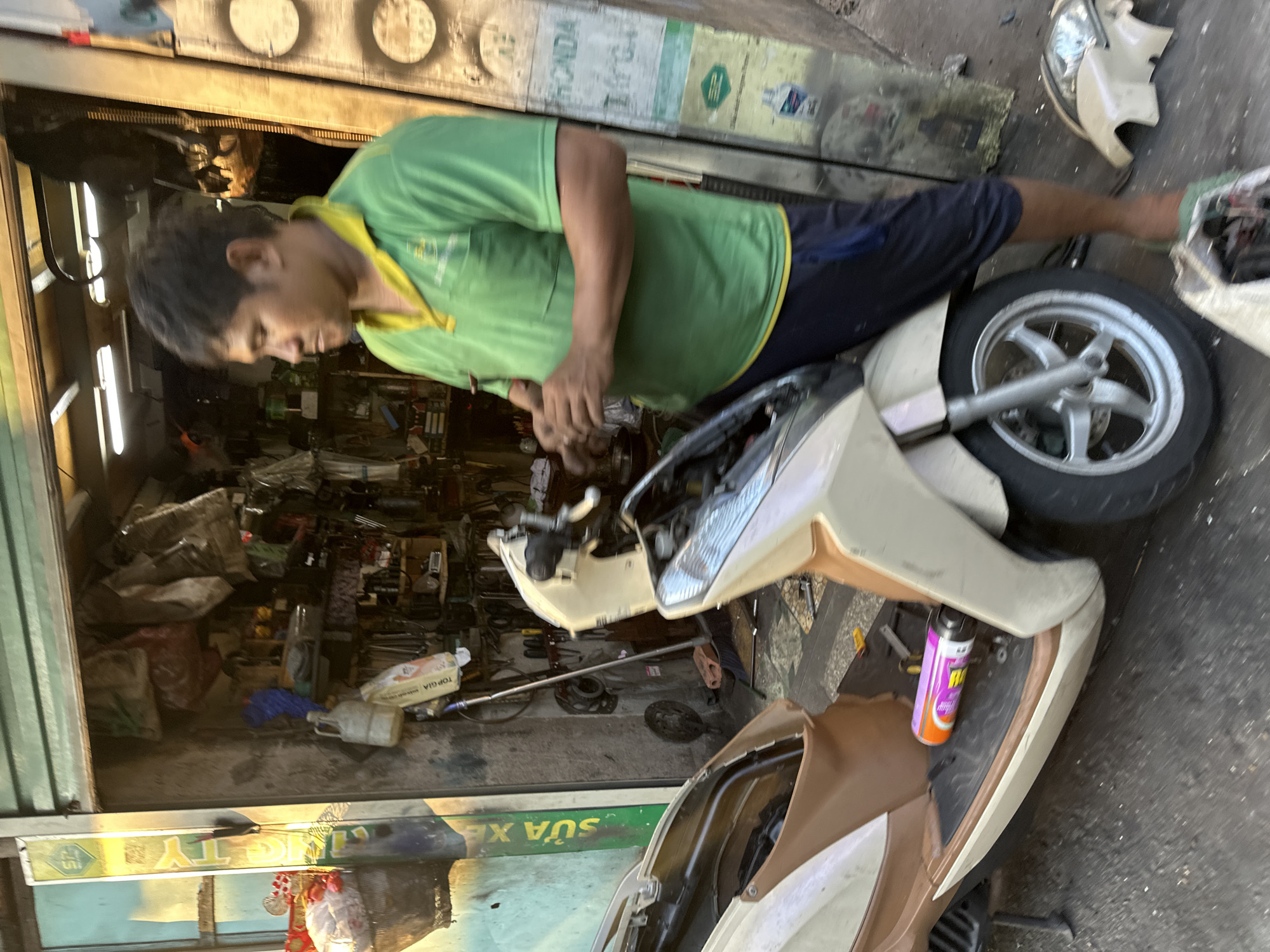 A man in a green and yellow t-shirt is working on a moped at a repair shop. The moped is light beige with brown accents. The front of the moped is disassembled, with the headlight lying nearby. Various tools and parts are scattered on the floor of the shop. The shop appears cluttered and disorganized. The sign above the shop front says "Sua Xe". The location is in Thủ Đức, Vietnam.