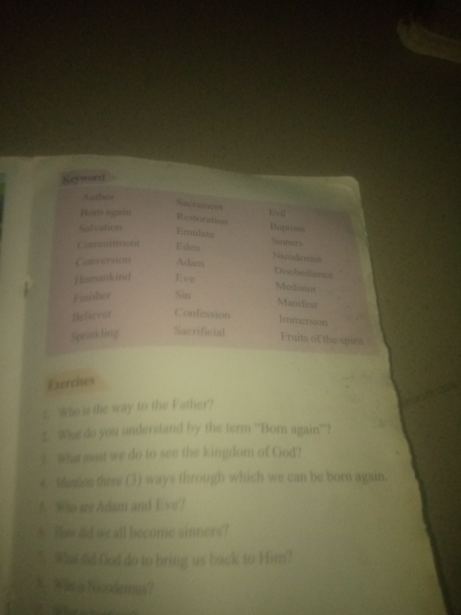 The image shows a close-up view of a page from a book, focusing on two columns of text. The left column, under the heading "Keyword," lists words like "Author," "Born again," "Salvation," "Commitment," "Conversion," "Humankind," "Finisher," "Believer," and "Sprinkling." The right column lists words such as "Sacrament," "Restoration," "Emulate," "Eden," "Adam," "Eve," "Sin," "Confession," "Sacrificial," "Evil," "Baptism," "Sinners," "Nicodemus," "Disobedience," "Mediator," "Manifest," "Immersion," and "Fruits of the spirit." Below these two columns, there is a section labeled "Exercises," followed by a numbered list of questions. The questions are: "Who is the way to the Father?", "What do you understand by the term 'Born again'?", "What must we do to see the kingdom of God?", "Mention three (3) ways through which we can be born again.", "Who are Adam and Eve?", "How did we all become sinners?", "What did God do to bring us back to Him?", and "Who is Nicodemus?". The lighting suggests the image was taken in a dimly lit environment, and the focus is on the printed text. The page appears slightly crumpled at the bottom right. The image does not contain any information about the city of Jauro Yinu, Nigeria, or any people.