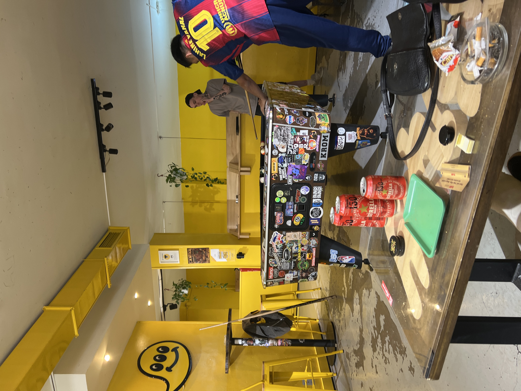 Here is a detailed description of the image:
**Scene:** The image appears to be taken inside a bar or pub. The interior design features extensive yellow accents, including walls, ventilation ducts, and furniture. A pool table covered in stickers is centrally located, with two individuals seemingly playing or preparing to play a game of pool.
**People:** Two people are visible. One is wearing a blue and red FC Barcelona jersey with the number 10 and the name "LAMINE VAMAL" on the back. He appears to be positioning himself to take a shot. The other individual, wearing a gray shirt and a black cap, stands near the pool table, possibly offering advice or waiting for his turn.
**Objects:**
- Pool table with stickers.
- Pool cue and other pool equipment on the table.
- Several cans of "Estrella Damm" beer and a green serving tray on a table.
- Ashtray with cigarette butts.
- A black purse.
- Wall-mounted lighting fixtures.
- Yellow ventilation ductwork.
- A smiley face with extra eyes on the wall.
**Actions:** People are engaged in the activity related to a pool game.
**Environment:** The bar or pub has a casual and relaxed atmosphere. The yellow color scheme contributes to a vibrant and energetic ambiance.
**Location:** There are two clues in the image that the location of the scene is in the city of Barcelona, Spain:
1. The FC Barcelona jersey worn by the individual in the photo is a professional association football club based in Barcelona.
2. "Estrella Damm" beer is a well-known beer from Barcelona.