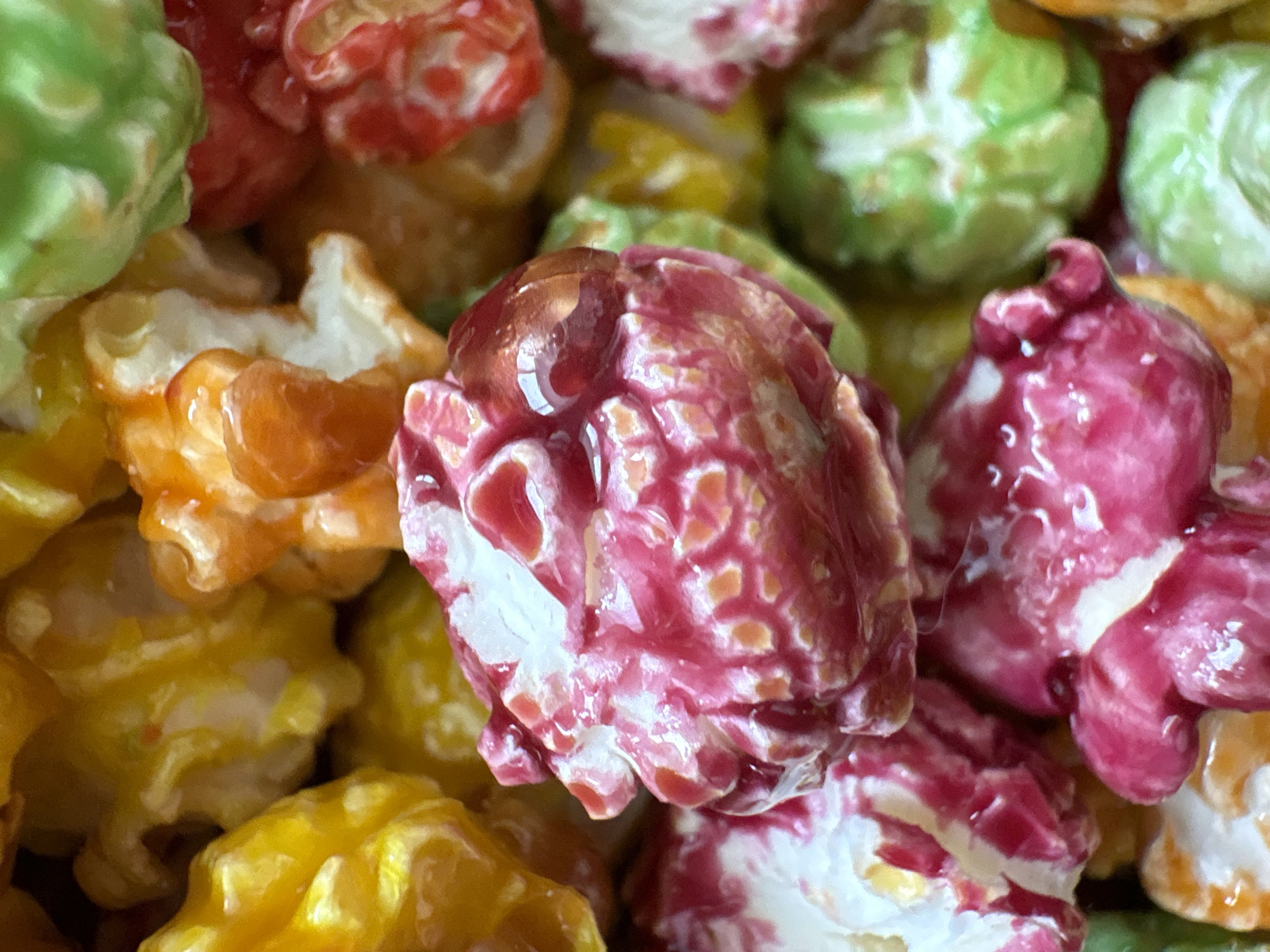 The image shows a close-up of candied popcorn in various colors: green, red, yellow, orange, and pink. The popcorn is coated with a shiny, glossy glaze. The scene depicts a collection of candied popcorn, not indicative of a specific location in Chaikovsky, Russia.
