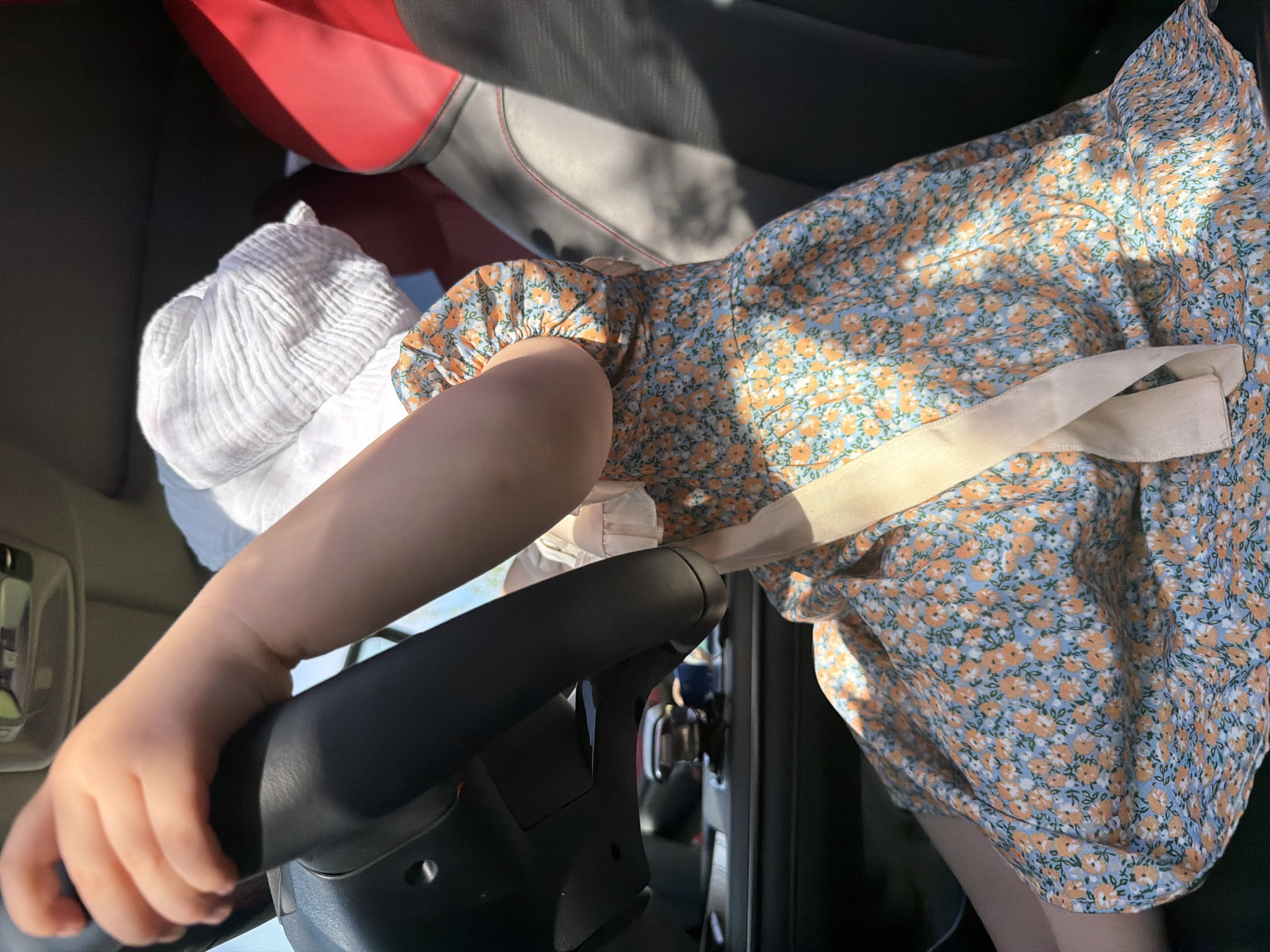 A child in a blue and orange floral dress and white sun hat is sitting in the driver's seat of a car. The child's left hand is gripping the steering wheel. The interior of the car, including the seats and dashboard, are visible. The specific location within Chaikovsky, Russia, cannot be determined from this image.