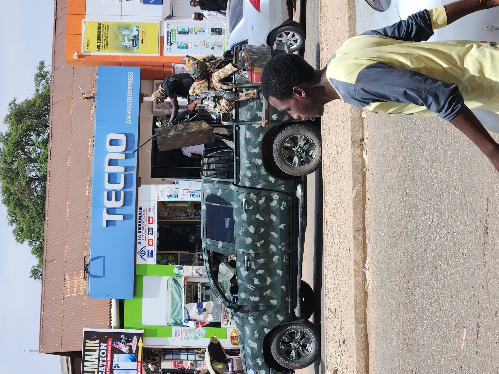 The image depicts a street scene in an urban environment of Unknown, Unknown, with a focus on a camouflage-painted pickup truck parked next to a sidewalk. In the truck bed, there are armed soldiers in camouflage gear, one holding a rifle. A man wearing a yellow and grey shirt is standing on the sidewalk facing the truck. Behind the truck, there are several storefronts. Prominently displayed is a large "TECNO" sign, indicating a mobile phone store, along with signs for "ZAKMAN ENTERPRISES" and "HAMAZ COMMUNICATIONS." There is also a banner for "LMALIK COMMUNICATION." Other pedestrians and vehicles are visible in the background.