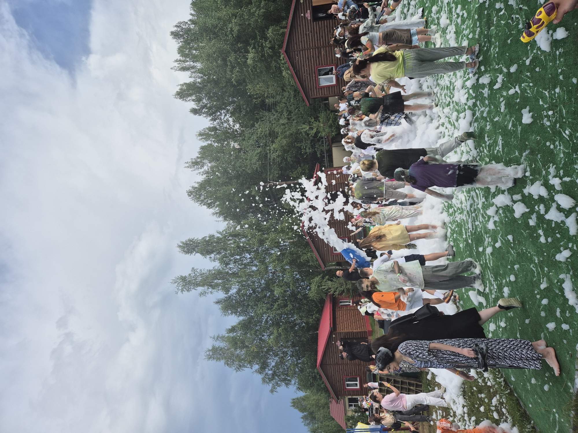 A group of people are gathered outdoors, on a bright green artificial turf surface, covered in a thick layer of white foam. They are participating in a foam party. In the background, there are several rustic wooden cabins with red roofs, and a dense line of tall green trees. The sky is overcast with some patches of blue. A man in the foreground is holding a yellow and purple water gun.
