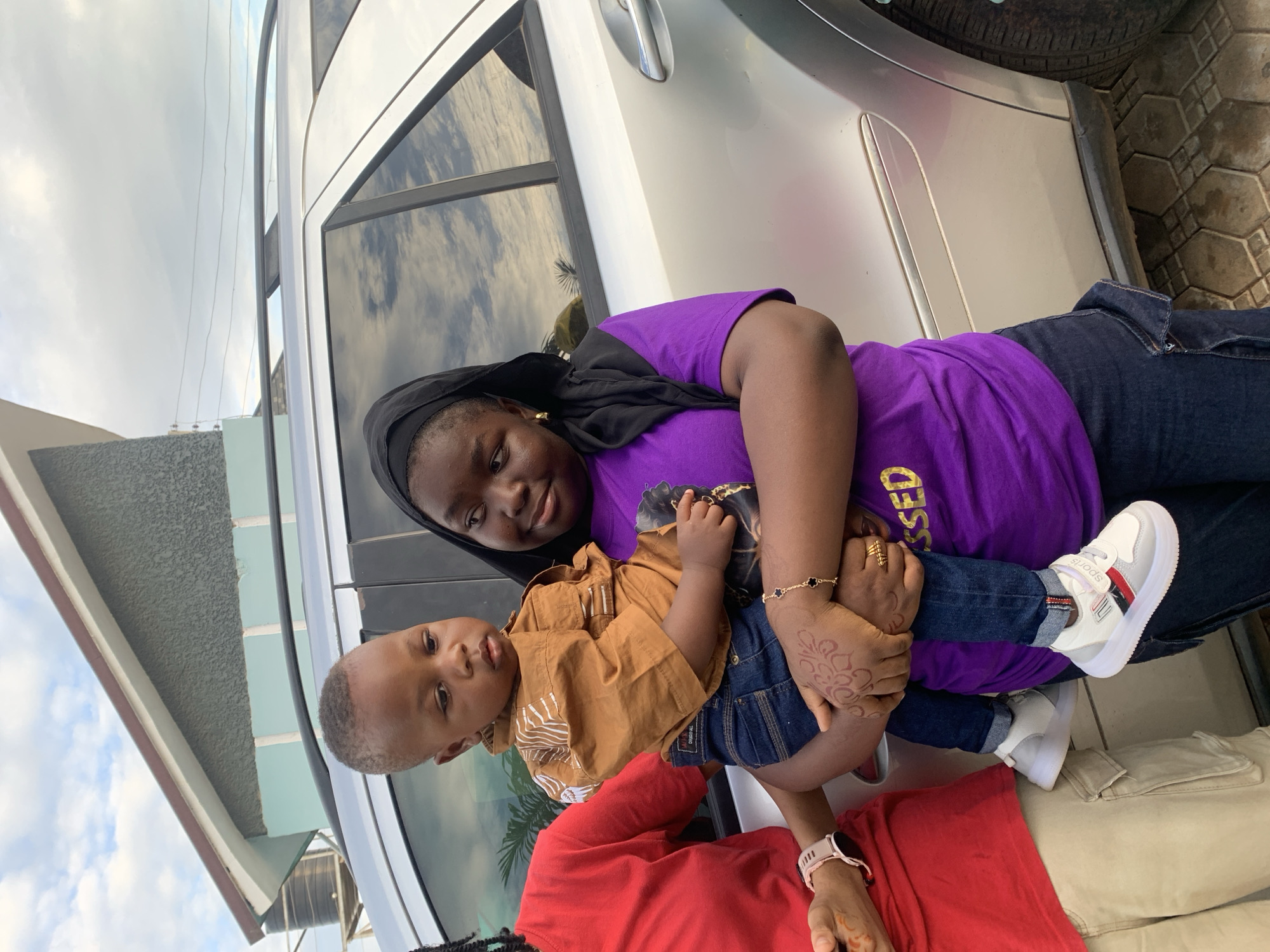 An outdoor, daytime scene in Jos, Nigeria, depicts three individuals next to a parked silver SUV. A woman on the right, wearing a black head covering, a purple t-shirt with partial yellow text, and dark blue jeans, holds a young male child. She has gold hoop earrings, a gold bracelet with black clover, and gold rings on her right hand, which also displays a henna design. Her gaze is directed at the child with a slight smile. The child, wearing a light brown collared shirt with white patterns, dark blue jeans, and white sneakers with "SPORTS" branding, looks downwards and to the left with a neutral expression. Partially visible on the left is another individual wearing a red long-sleeved shirt, tan cargo pants, and a light-colored smartwatch on their left wrist; their left hand also shows a henna design and is extended towards the center without direct contact. Behind the individuals, a silver SUV, possibly a Mercedes-Benz, features a black roof rack and reflective windows showing cloud patterns. In the upper left background, a building with a grey textured upper facade, a light blue lower section, and a brown roof trim is visible, with a black water tank on a stand situated behind it. The ground is paved with hexagonal interlocking tiles. The sky above is partly cloudy.