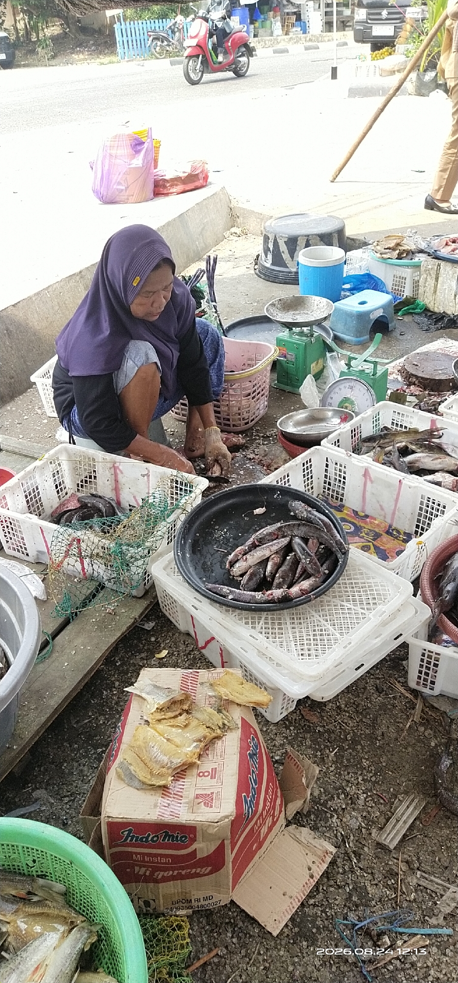 In Tamiang Layang, Indonesia, an outdoor scene depicts a woman wearing a purple hijab and brown gloves, crouched down, cleaning fish. She is surrounded by various containers and objects related to fish sales. To her left, a large green plastic basket holds numerous small fish. In front of her, a white plastic crate also contains fish, some covered by a green net. Adjacent to this crate is a black circular tray filled with more fish. Behind the woman, a pink woven basket holds fish. Several white plastic crates, some stacked, are visible around the area, and in the background, various containers, scales, and a blue stool are present. To the right of the woman, a person in beige pants and black shoes stands holding a long wooden stick. The ground is covered with gravel and debris, and a cardboard box with "Indomie Mi Goreng" branding is on the ground, topped with dried fish. In the distance, a red scooter is parked near a blue fence, and a paved road with vehicles is visible.