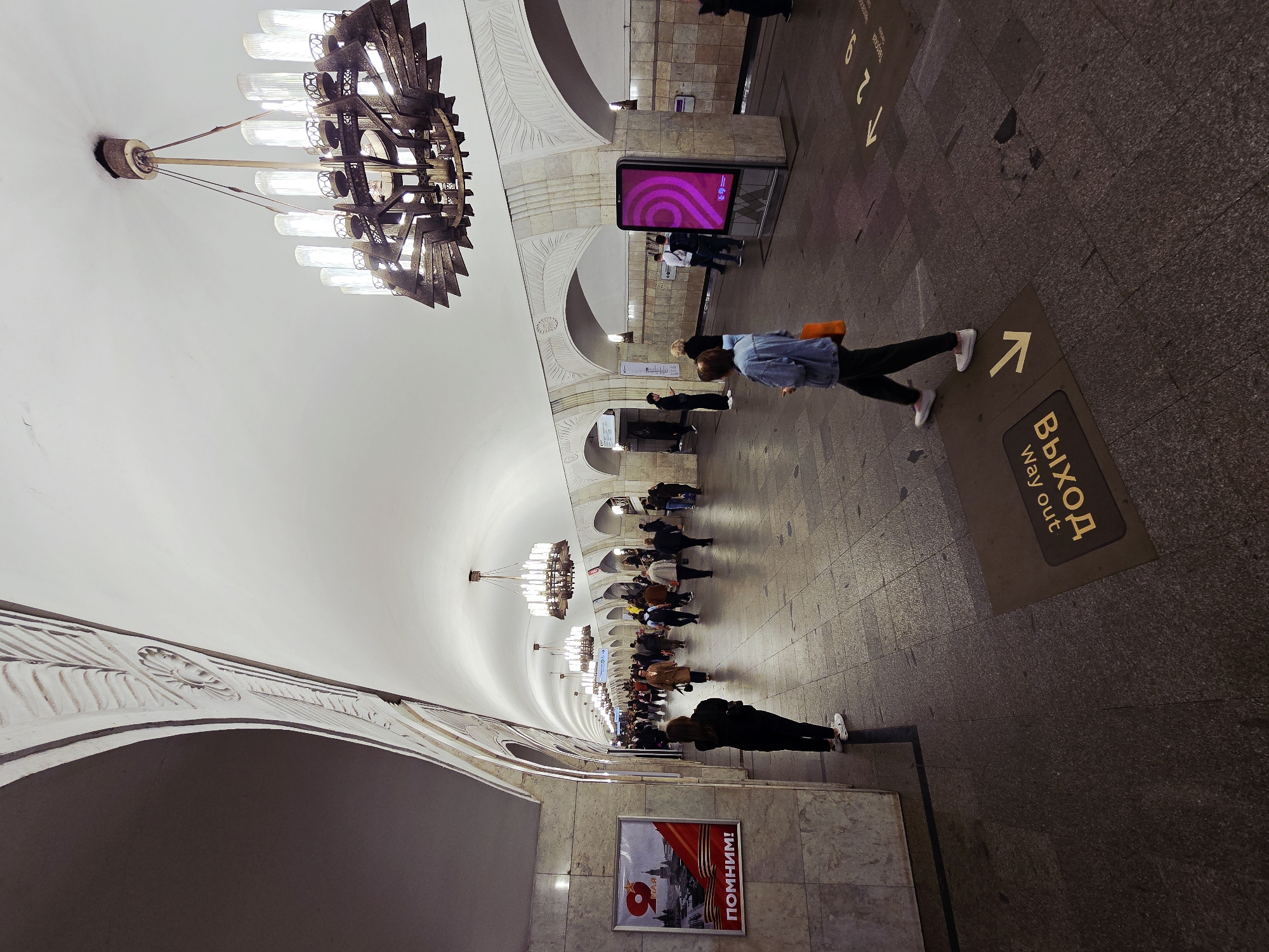This image shows a corridor inside a Moscow Metro station in Russia. The station is characterized by its high, vaulted ceiling adorned with ornate chandeliers and decorative arches. People are walking along the tiled floor. There's a sign on the floor with an arrow. Along the walls, there are advertisements and informational displays, and a poster. The overall scene depicts a busy transportation hub.