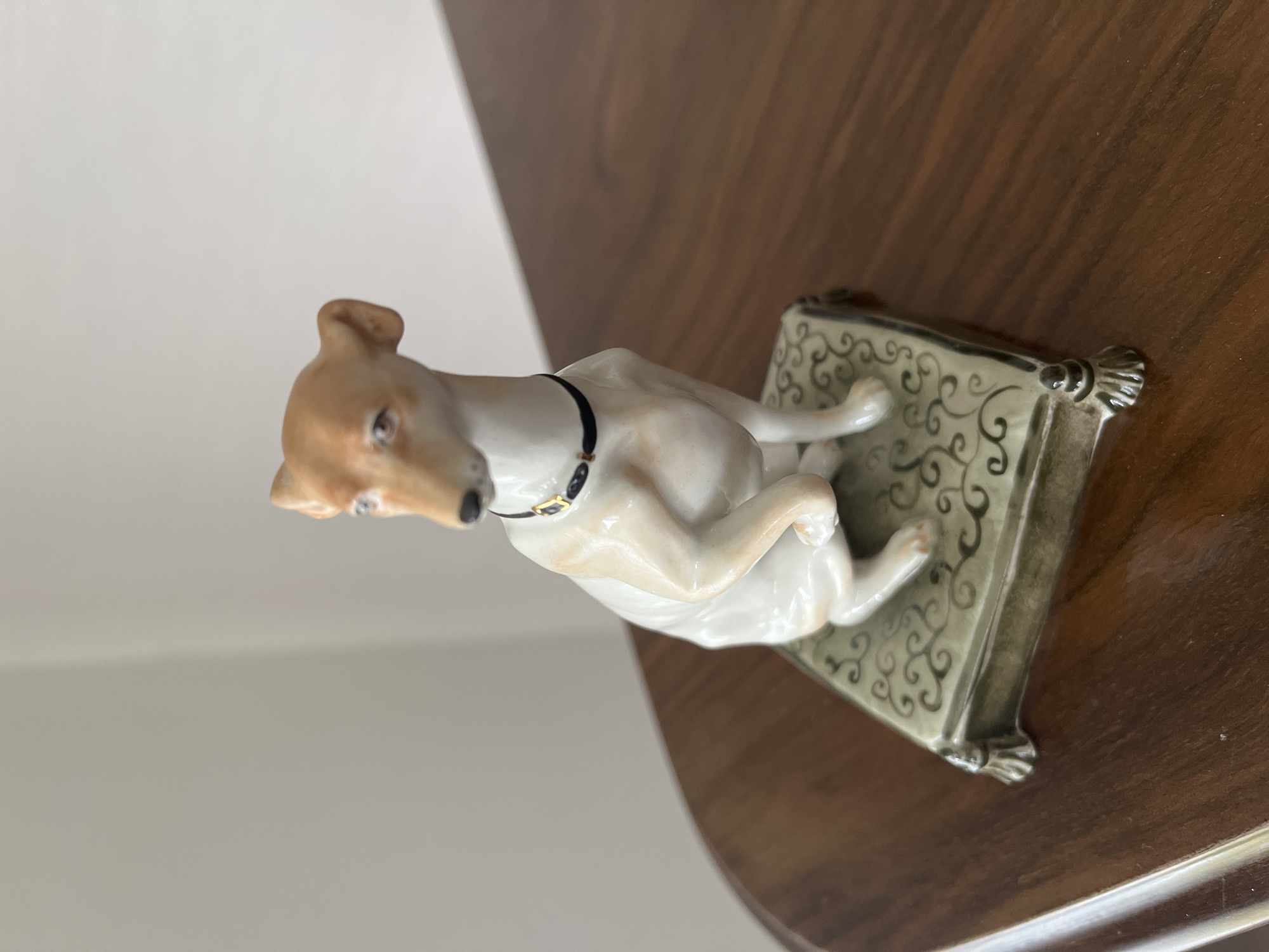 A porcelain figurine of a dog is perched on a decorative green base. The dog is white with tan markings on its head and body, and it wears a black collar with a gold accent. The base has an ornate pattern and four small, decorative feet. The figurine sits on a polished wooden surface, possibly a tabletop or dresser. The background is a plain, light-colored wall. The lighting is soft and natural.