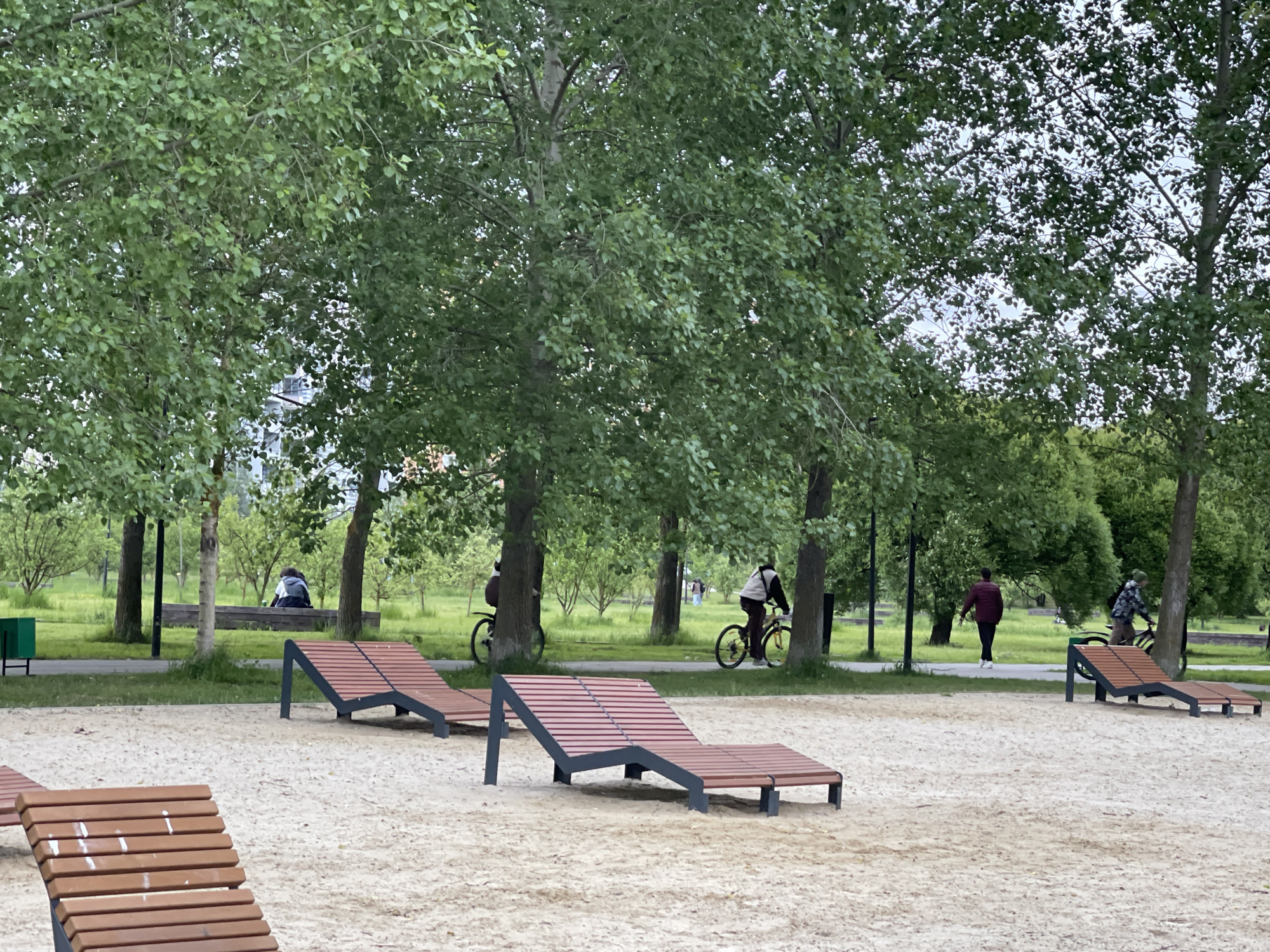 The image depicts a sandy area with several wooden lounge chairs. In the background, a paved path is visible, lined with trees. People are walking and cycling along the path. The location is in a park in the city of Moscow, Russia.