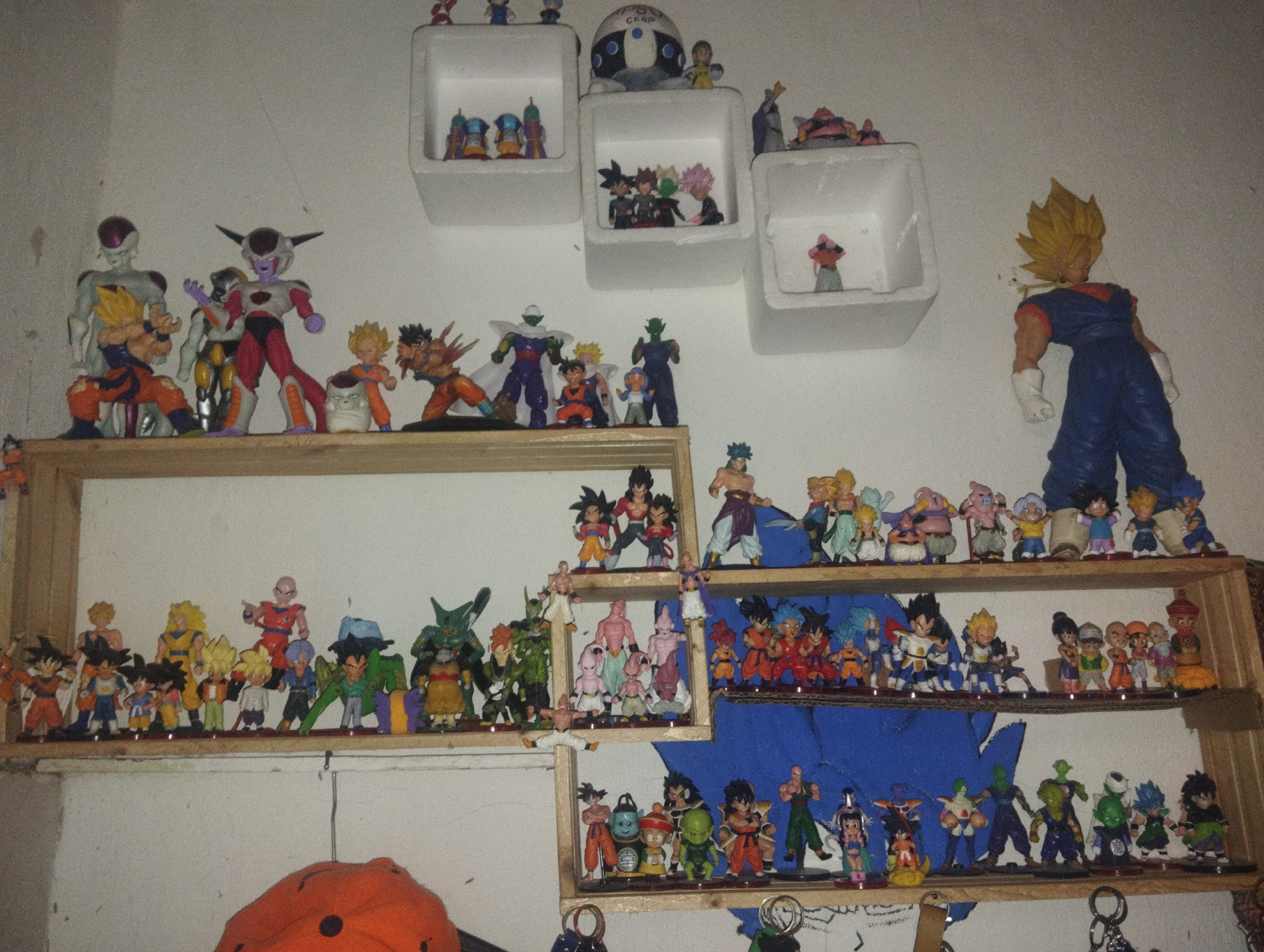The image shows a collection of Dragon Ball Z figurines displayed on shelves and in wall-mounted cube containers. The figurines depict various characters from the series, including Goku, Vegeta, Frieza, Piccolo, and others in different forms and stages of transformation. The shelves are made of light-colored wood, and the cube containers are white. The wall is painted a light color. There is a blue backdrop behind some of the shelves. The location is El Valle del Espíritu Santo, Venezuela. There are no people visible in the image.