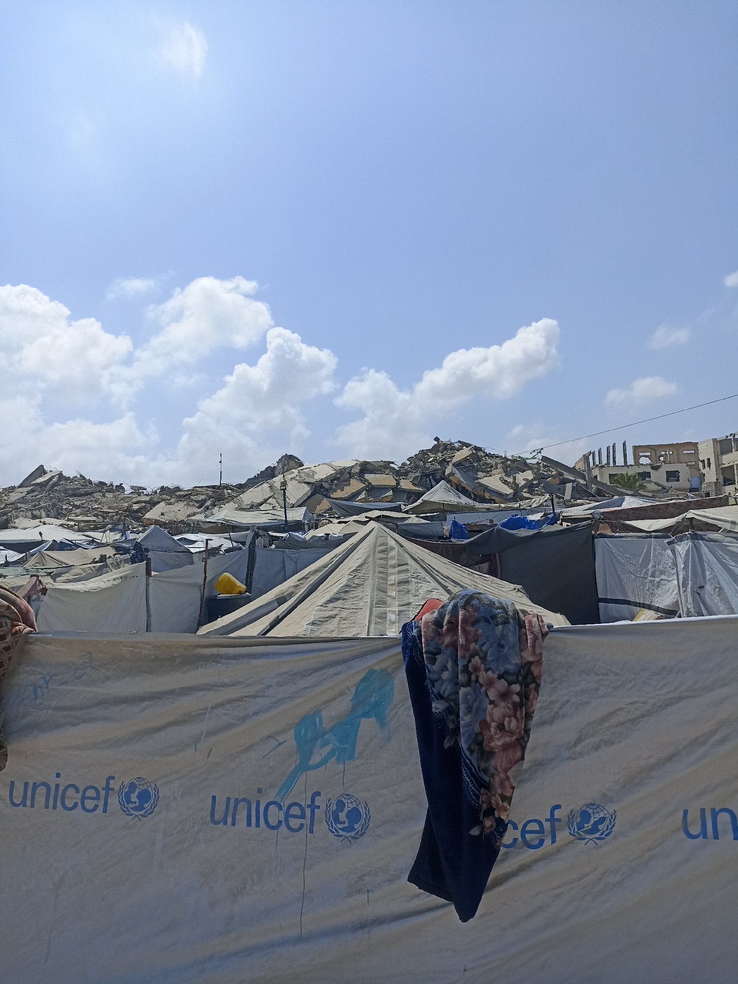 In Jabalia, Palestinian Territories, a tent city has been established amidst the rubble of destroyed buildings. The foreground features a large white tarp with blue UNICEF logos. Two blankets, one floral and one dark blue, hang over a clothesline or barrier in front of the tarp. Beyond the tarp, numerous tents of various sizes and colors are erected, forming a dense encampment. In the background, the devastating aftermath of destruction is evident with large piles of concrete debris and damaged structures. The sky is blue with scattered white clouds.