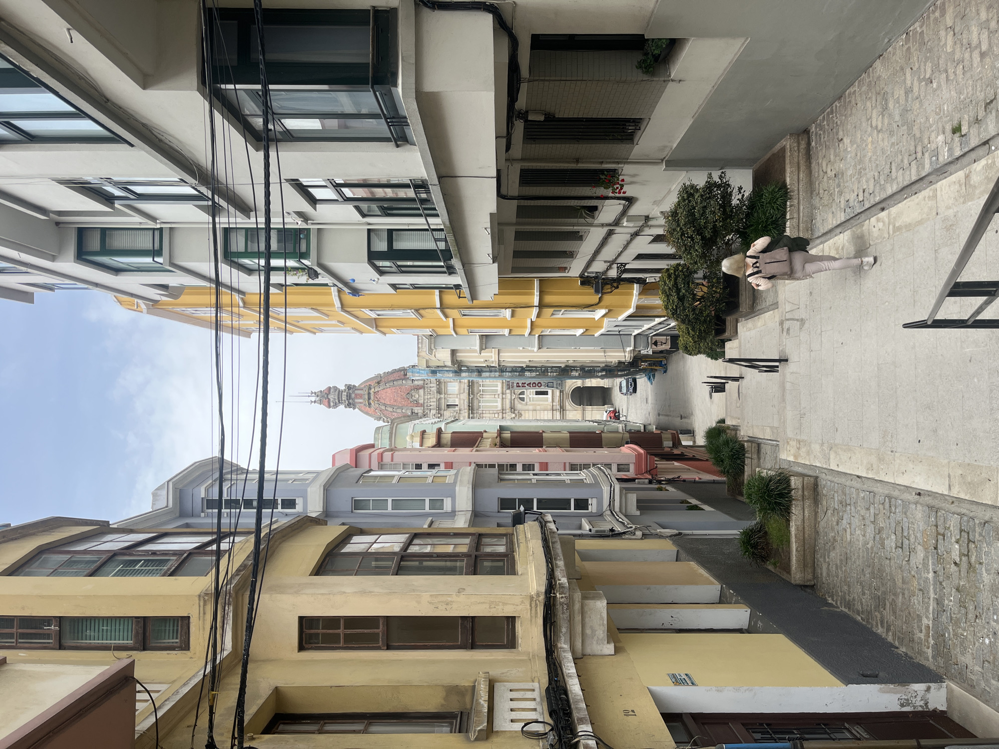 A downward-angled, medium-distance shot reveals a street scene in A Coruña, Spain. The perspective is drawn along a narrow pathway between tall, colorful buildings leading toward a distant structure with a pink dome. A single person, walking away from the viewer, ascends a set of steps on the right side of the frame. The buildings are primarily in pastel shades of yellow, grey, and pink. Electrical wires are visible crisscrossing the space between the buildings. Vegetation is present along the pathway. The sign "PRADO" is partially visible at the end of the street, indicating the direction. A parked car is also visible near the end of the street. The sky is overcast and appears to be a light blue-grey.
