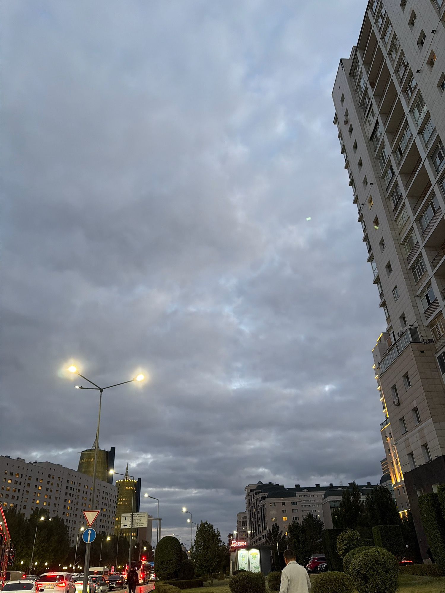 The image captures a street scene in Astana, Kazakhstan, on an overcast day with heavy cloud cover. The perspective is from street level, looking upwards towards buildings and the sky.
On the right side of the frame, a tall, modern apartment building dominates the view. Its facade is light-colored with numerous balconies and windows, some of which are lit from within. The building's structure is angled and geometric, with architectural details illuminated by warm exterior lights.
To the left, the sky is filled with dark, moody clouds. A street lamp with three lights, two of which are brightly illuminated, stands tall, casting a warm glow. In the mid-ground, a busy street is visible with a line of cars, indicating traffic flow. Several vehicles are in motion, with their headlights and taillights visible. Traffic lights and road signs are present, including a blue circular sign with a white arrow pointing upwards and a red and white triangular sign.
Further in the background, other buildings are visible. One has a distinctive, pointed architectural design and is lit up, suggesting it's a prominent structure. Another, wider building to the left is also illuminated with many windows. Trees and manicured bushes line the street and sidewalks, creating a sense of urban landscaping.
In the foreground, a person wearing a light-colored jacket is walking away from the camera on a paved area. Another person, partially obscured, is visible near a bus with its lights on. There are some kiosks or small structures near the sidewalk, one of which appears to have displays of flowers. The overall impression is of a dynamic urban environment under a dramatic sky.