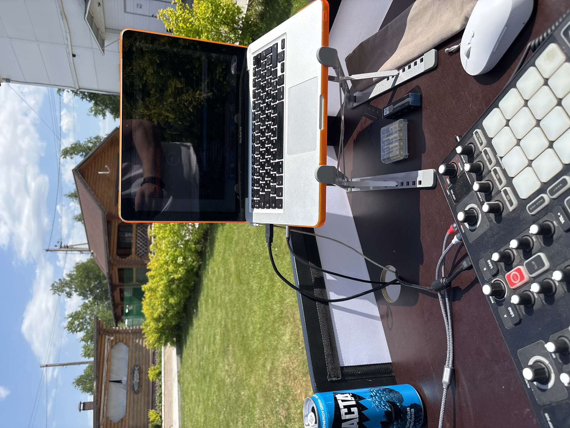 Observed at an outdoor setting in what appears to be Chaikovsky, Russia, a laptop computer with an orange case sits on a stand on a brown table, a musical instrument next to a white mouse. Also on the table are a USB flash drive, a battery case, and a can of blue ACTA Energy Drink. In the background is a wooden house. The sky is partly sunny.