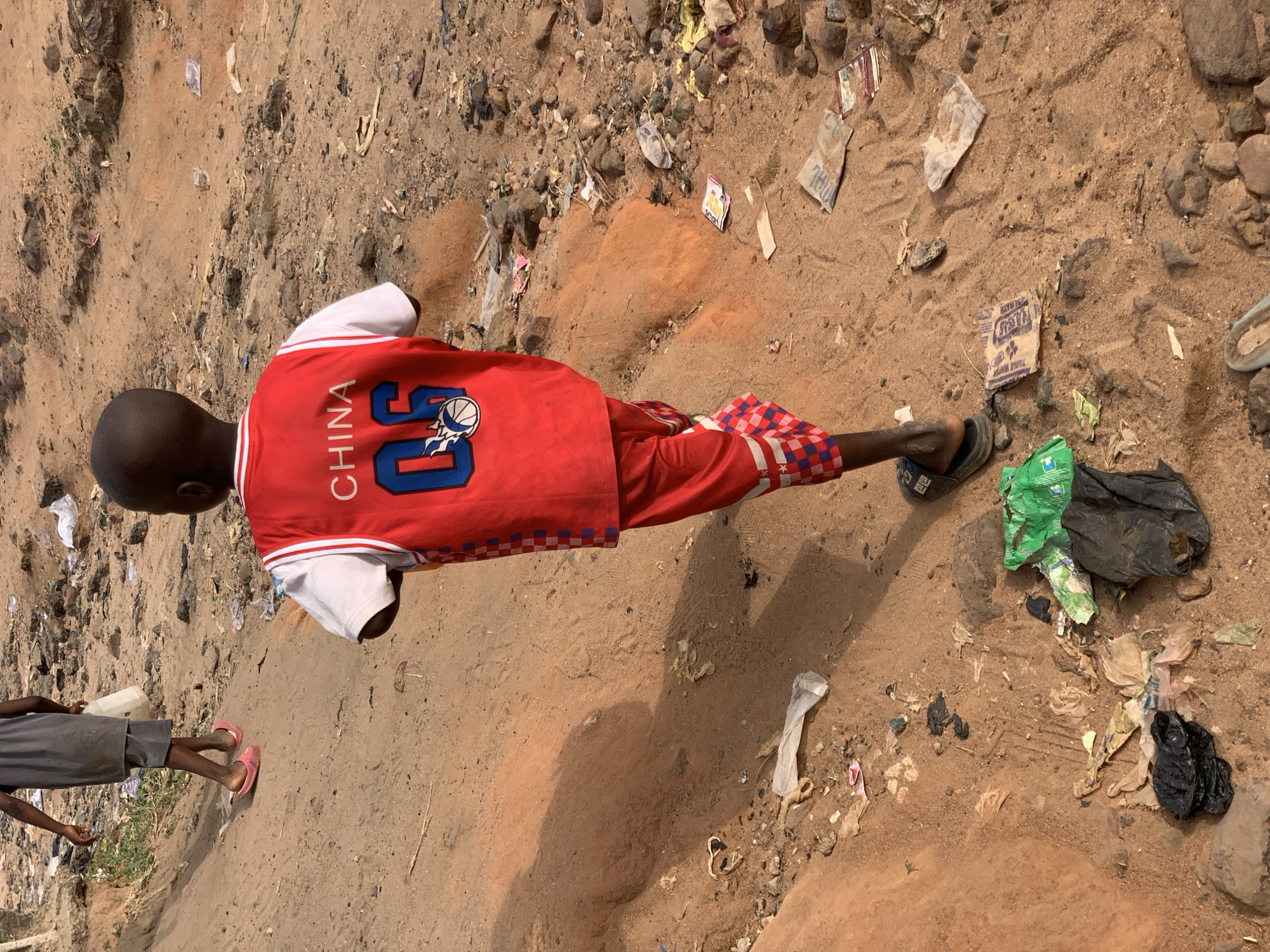 The image captures an overhead view of an outdoor scene in Lokoja, Nigeria, featuring at least two individuals on a sandy, reddish-brown dirt path. The ground is uneven and extensively littered with various items such as plastic bags (green, black, and clear), paper wrappers, and other debris.
The primary subject is a young person, likely a child, with dark skin and a bald head, seen from behind. They are wearing a vibrant red and white basketball-style jersey with red checkered shorts and dark sandals. The back of the jersey prominently displays the word "CHINA" written vertically in white letters, alongside the number "05" and a stylized basketball logo with white flames and blue outlines.
A second person is partially visible on the left side of the frame, wearing grey attire and pink flip-flops, standing near a white container. Both individuals appear to be standing casually, with no specific activity discernible.
The bright, direct lighting and the distinct shadows cast on the ground indicate it is daytime, likely under a clear sky. The setting appears to be an unpaved, possibly informal or undeveloped area.