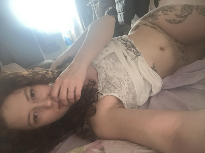 The specific location within the city of Saint Petersburg, Russia, cannot be determined from the image provided.
The scene depicts a female individual with dark, curly hair, lying on her left side in an indoor setting. She is positioned on a bed covered with light-colored bedding, which appears to be purple in some areas and has a floral pattern in others. The individual is wearing a light-colored, patterned top that reveals her midriff. Extensive tattoos, featuring botanical or floral designs, are visible on her upper left thigh and hip. A simple silver-colored ring is present on her right ring finger. Her right hand is raised towards her mouth, with fingers partially obscuring her lower face. Her gaze is directed towards the camera. The background on the left side of the frame is overexposed due to a bright light source, likely a window. On the right side of the background, indistinct objects and dark fabric, possibly clothing, are partially visible.