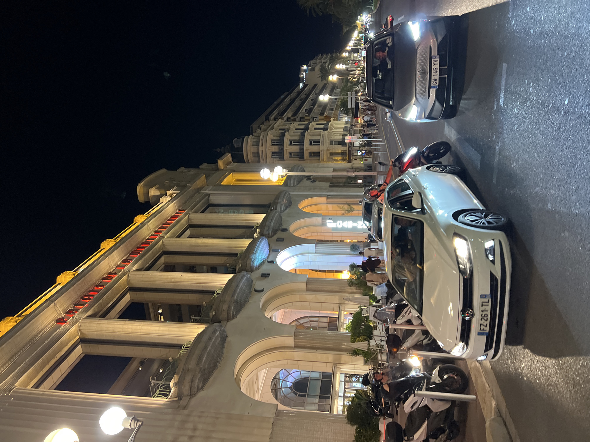 At night, on a city street in Nice, France, a white Volkswagen Polo with license plate FZ-261-TL is in motion, its headlights illuminated. To its right, a silver MG ZS EV with license plate GW-146-YV is also in motion, its headlights on. Several parked scooters are visible on the left side of the street, with a person on one of them wearing a helmet. In the background, a large, ornate building with arched entrances and classical columns is illuminated. People are walking on the sidewalk in front of the building, and further down the street, several multi-story buildings line the road. The scene is illuminated by streetlights and the lights from the buildings.