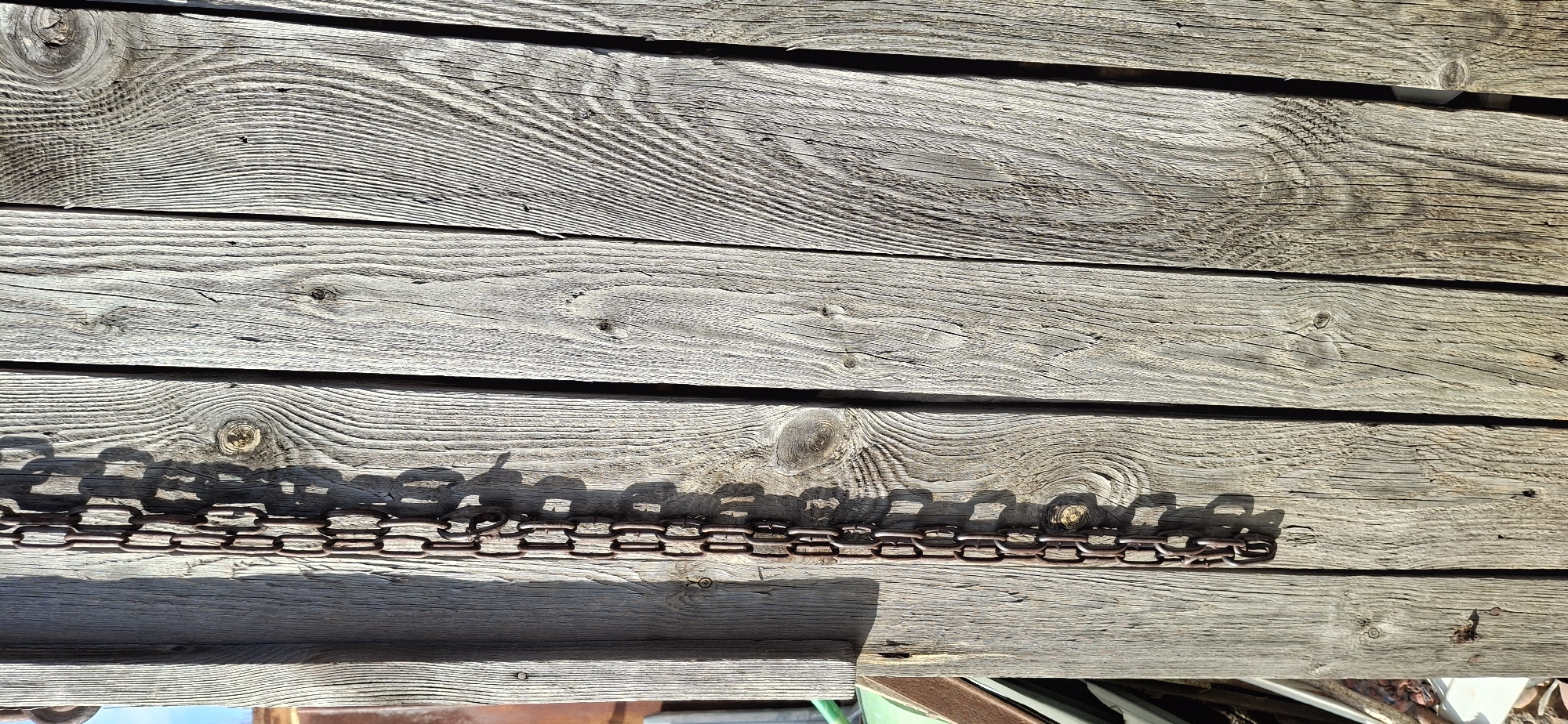 An eye-level, outdoor shot depicts a weathered wooden structure, potentially a fence or wall, consisting of horizontal planks of varying widths. The wood exhibits prominent grain patterns and knots, indicative of age and exposure to the elements. A metal chain lies along one of the planks, casting shadows on the surface. The chain appears to be made of small, interconnected links. Other objects and details are partially visible at the bottom of the frame. The location is presumed to be in Chaikovsky, Russia, based on the user's query, but this cannot be confirmed visually from the image alone.