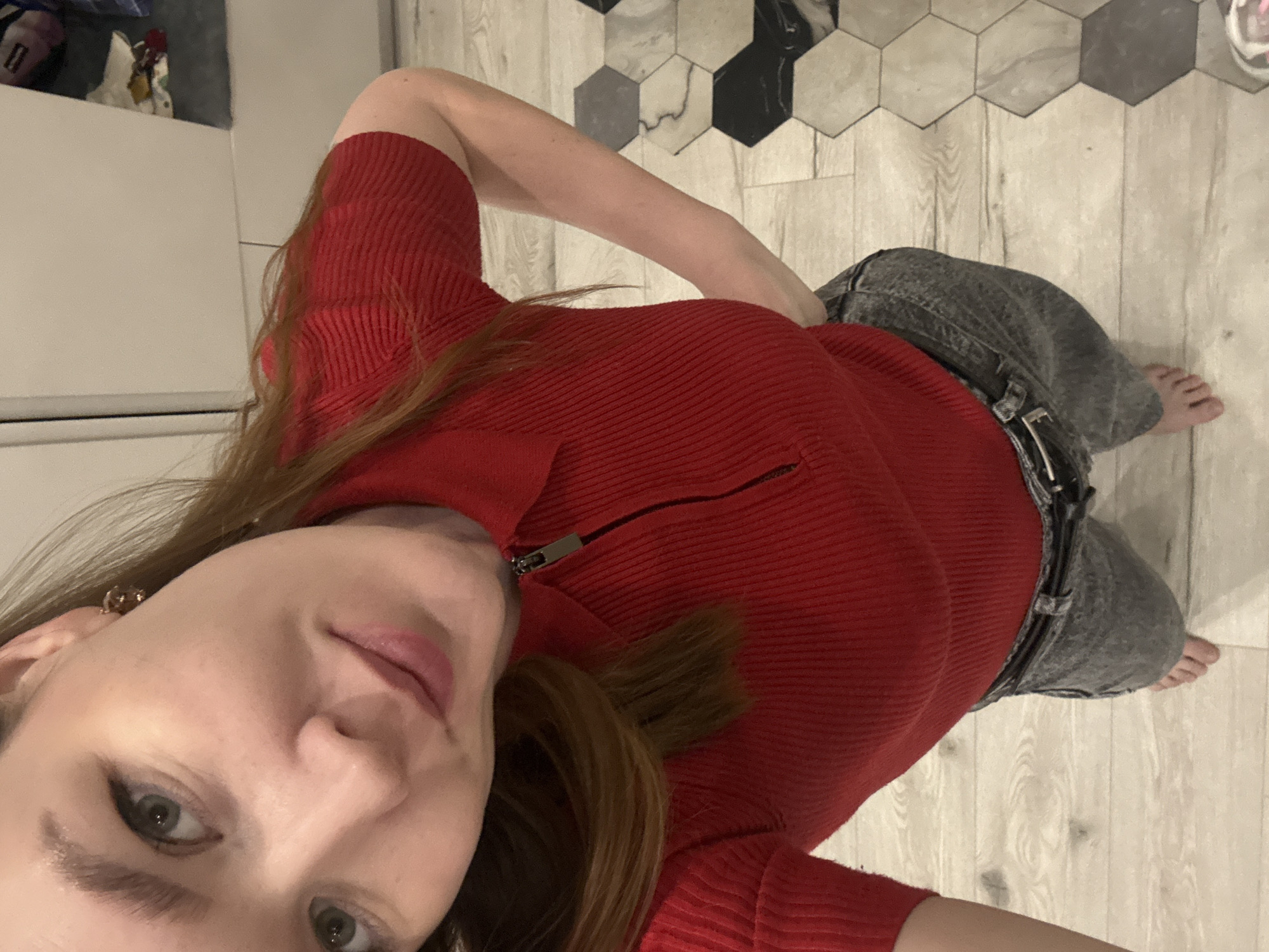 A young woman stands indoors, facing the camera from a high angle. She wears a red, ribbed, long-sleeved top with a partially zipped front and gray denim pants with a black belt. Her hair is brown. She is barefoot. The background includes light-colored wood-look flooring, some with decorative black and gray hexagonal tiles. Part of a white cabinet is visible in the upper left corner. There is no specific information to indicate that the photo was taken in Izhevsk, Russia.