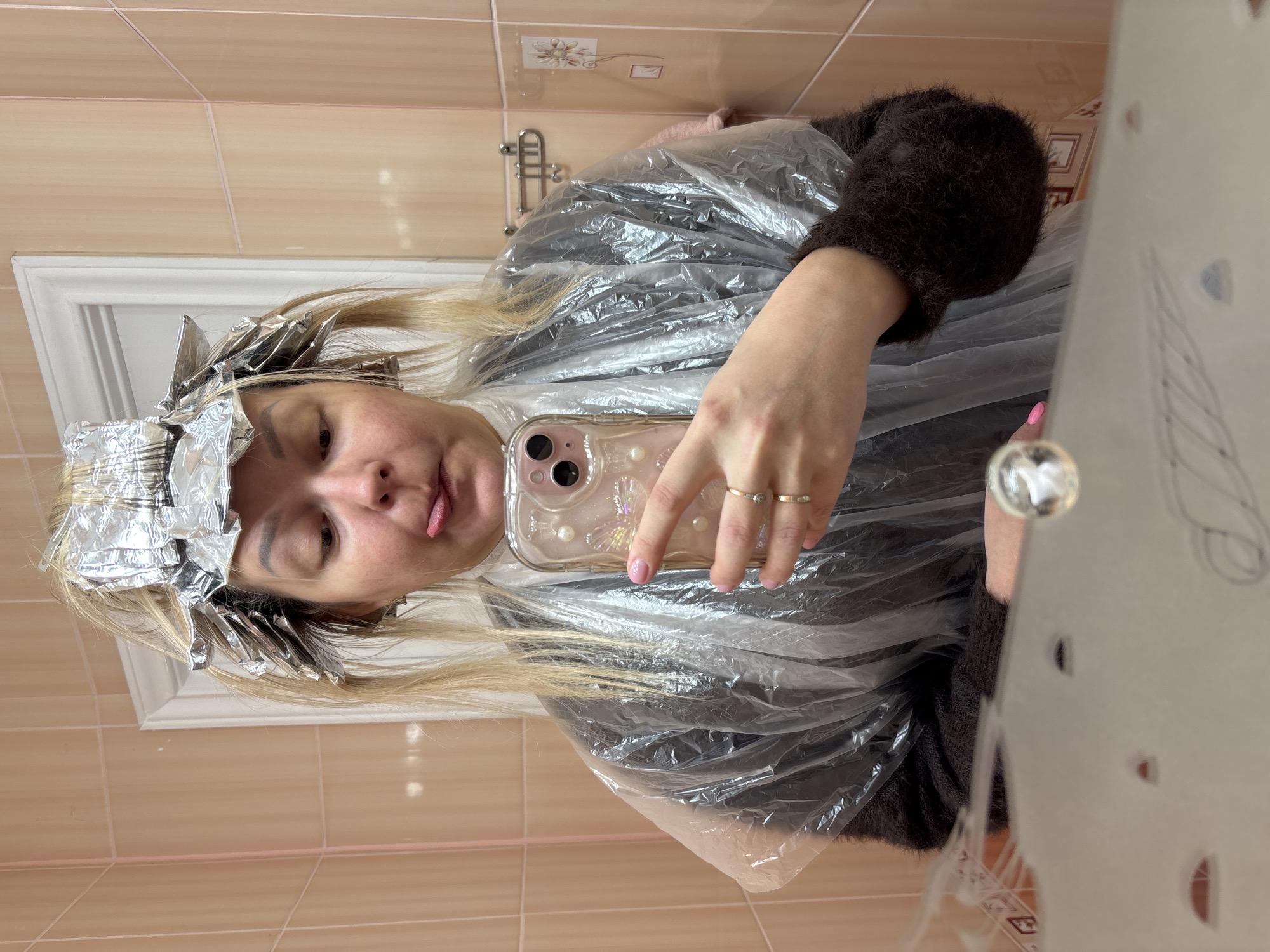 A woman is taking a selfie in a bathroom. She has blonde hair with foil highlights being applied, a plastic cape over a brown fuzzy top, and is holding a cell phone with a pearlescent case. The background includes tan tiled walls and a chrome faucet. The scene appears to be a home bathroom. The city and country are not discernible from the image.