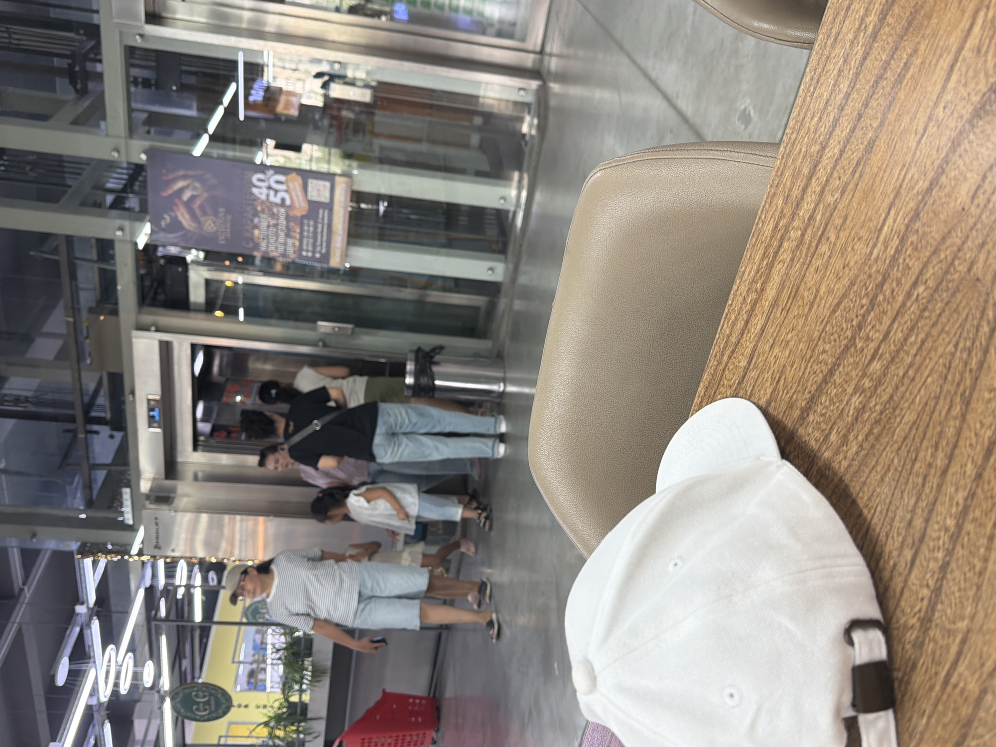 The image shows a ground-level view of a modern indoor space, likely a shopping mall or a similar public venue. In the foreground, a wooden table is visible, with a white baseball cap resting on it. The cap is white with a dark buckle on the strap. The table has a prominent wood grain pattern.
In the mid-ground, a group of people are standing near an elevator bank. One woman, wearing a striped t-shirt, a beige cap, and light blue shorts, is looking towards the left. To her right, a young girl in a white top and light blue shorts stands. Behind them, another woman in a black t-shirt and light blue jeans is visible, as well as other individuals, their faces mostly obscured. The elevator doors are made of glass, revealing the interior of the elevator shafts.
In the background, there are signs and bright overhead lighting. One sign, partially obscured by reflections, appears to be advertising a sale with percentages like "40" and "50". The environment has a polished concrete floor and glass and metal architectural elements. To the left, a red shopping basket is partially visible. The overall impression is a busy, well-lit commercial area.