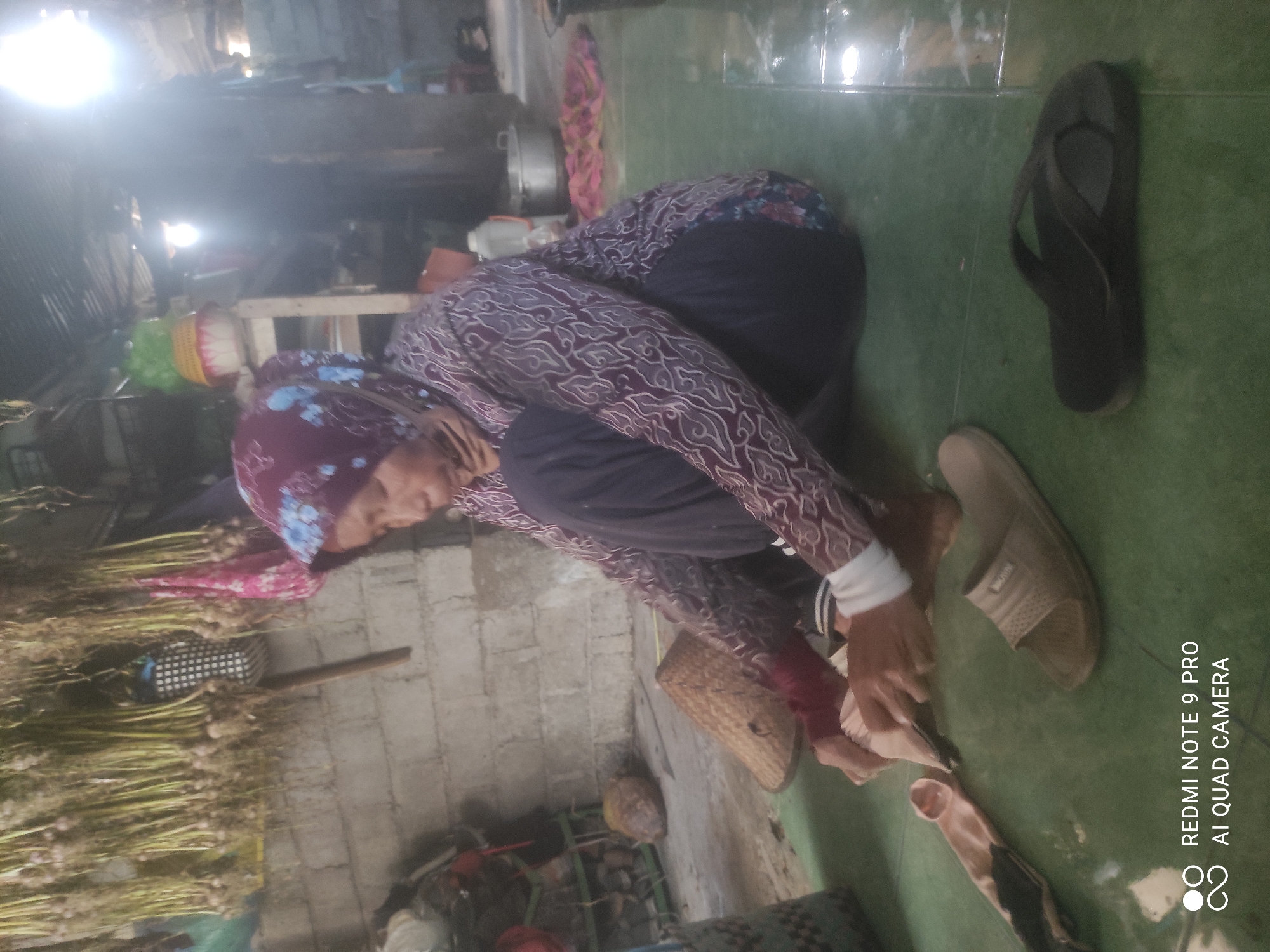 An elderly woman is seated on a green tiled floor, wearing a maroon hijab with a floral pattern and a patterned long-sleeved shirt. She is looking down, working with her hands, which appear to be holding a light-colored object, possibly fabric or food items. Her legs are crossed, and she is wearing dark pants. In front of her, to her right, are two pairs of footwear: a tan sandal and a black flip-flop. To her left, a woven conical hat is placed on the floor, and a light-colored sock lies nearby. The background depicts a rustic interior with a brick wall and items hanging from the ceiling, including what appears to be dried garlic or similar produce. Shelving units with various objects are also visible. The overall lighting suggests an indoor setting with natural light contributing to the scene. The image also contains a watermark in the lower-left corner, reading "REDMI NOTE 9 PRO AI QUAD CAMERA".
