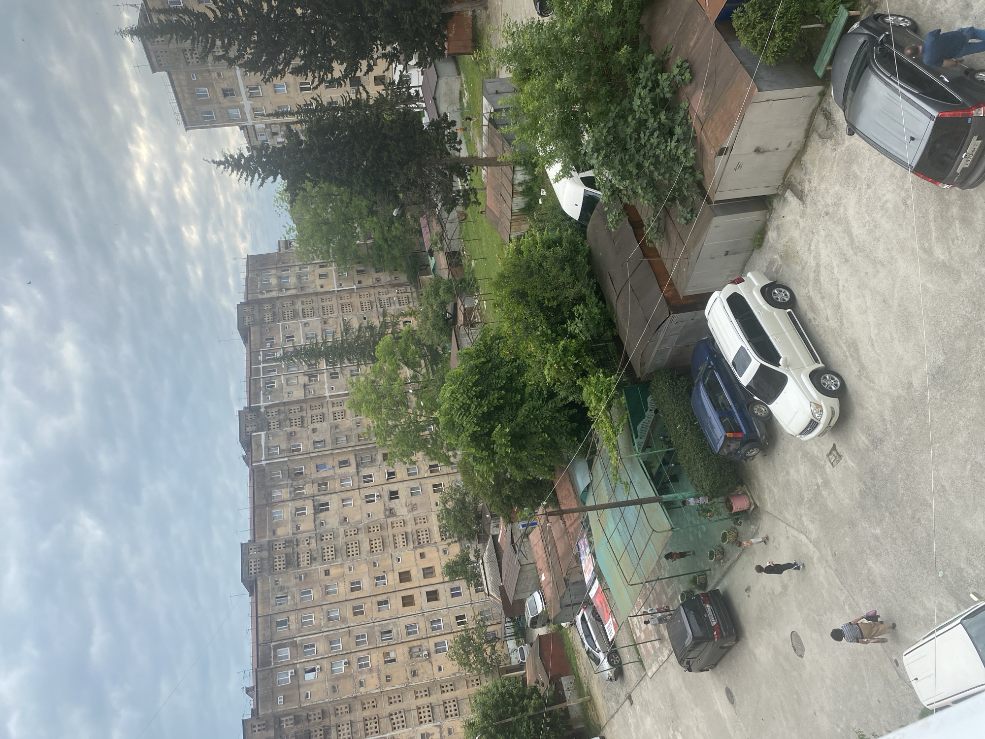 The image shows an aerial view of a courtyard in front of a Soviet-era apartment building. The building is multi-story with a repetitive facade of windows and what appear to be ventilation grilles. Several cars, including a black minivan, a blue SUV, and a white SUV, are parked in the courtyard. There are also several garages with corrugated metal roofs, and some green structures that may be covered seating areas or small market stalls. Green trees and bushes are scattered throughout the scene, and the sky is overcast. A few people are visible walking in the courtyard, including a woman carrying a bag and two children. One man is standing next to a black SUV. The overall impression is one of a lived-in urban environment.