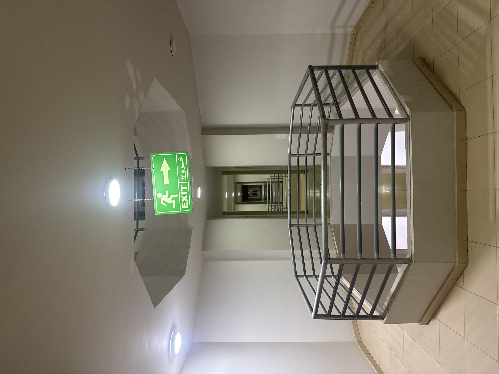 A low-angle shot presents a staircase extending into the distance within a building. The walls are white, and the floor has light tan tiles. A green "EXIT" sign with Arabic text hangs from the ceiling on the left side of the frame. A metallic railing extends from the right side of the frame toward the center, partially obstructing the view. Recessed lights are visible on the ceiling. The location is Unknown, Unknown.