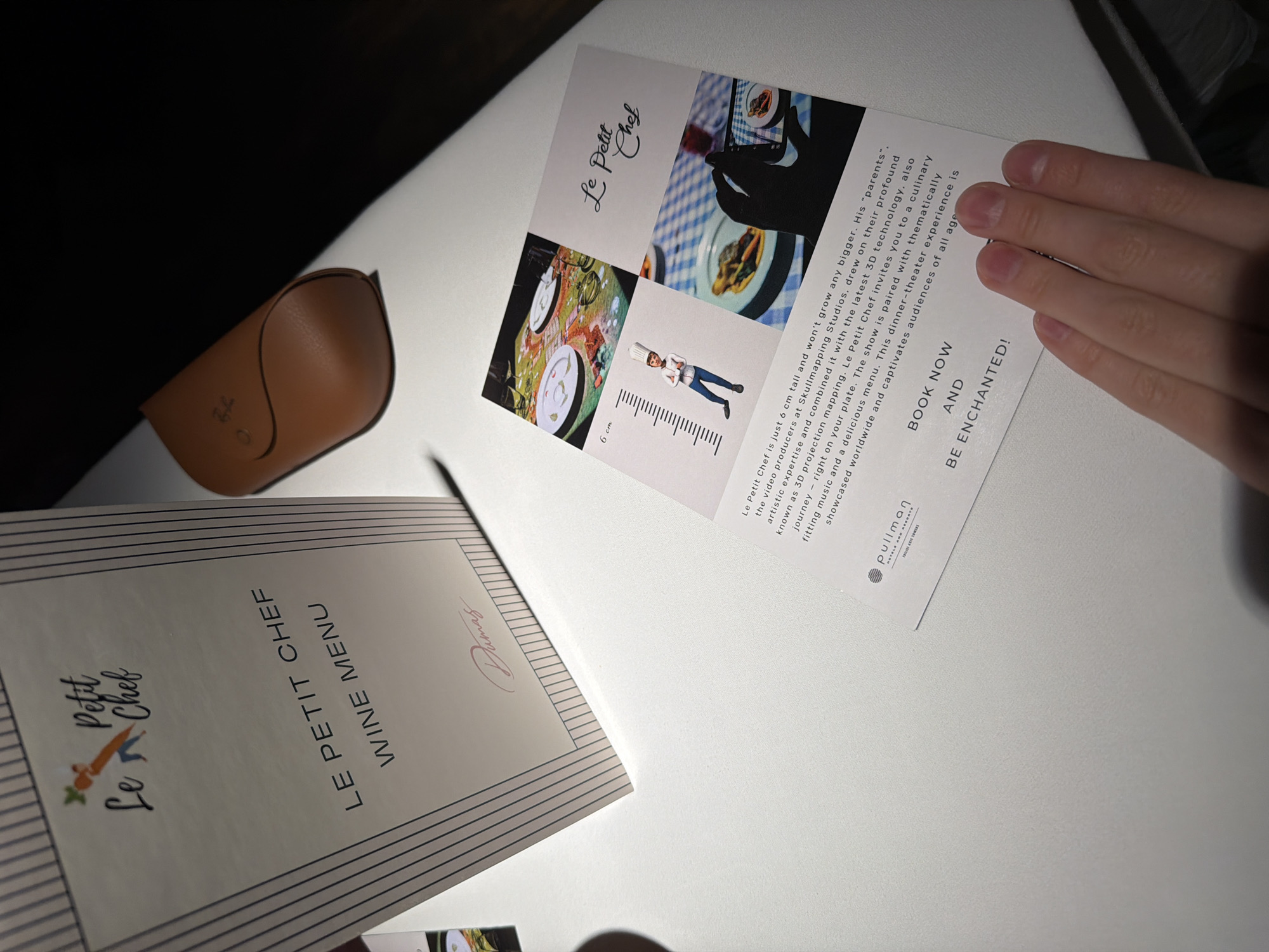 A close-up, top-down shot reveals a bright white table surface displaying promotional materials and a menu. A right hand, positioned on the right side of the frame, holds a white vertical card promoting "Le Petit Chef." Another white paper, titled "LE PETIT CHEF WINE MENU," lies flat on the left side of the table, featuring the "Le Petit Chef" logo and a signature "Dumas." A brown leather glasses case with a "Ray-Ban" logo is partially visible on the far left.
The card held by the hand describes "Le Petit Chef" as a 6 cm tall character created by "Skullmapping Studios" using 3D projection mapping, offering a culinary dinner-theater experience. It mentions a "culinary journey right on your plate" paired with "thematically fitting music and a delicious menu." The card features several images, including projected food on dining tables and the small chef illustration next to a ruler indicating "6 cm." At the bottom, it reads "BOOK NOW AND BE ENCHANTED!" and displays the "pullman HOTEL AND RESORTS" logo. The scene takes place indoors, likely at a hotel in Tbilisi, Georgia, under bright artificial lighting.