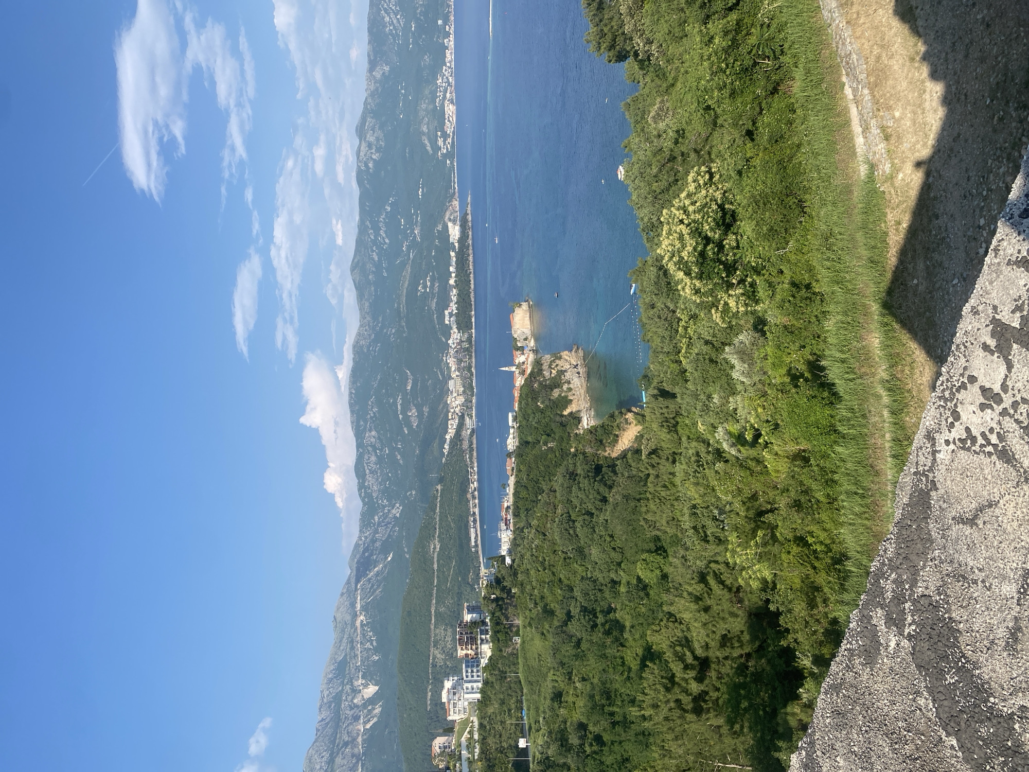 From an elevated position, the image looks over the coastal region of Budva, Montenegro. Mountains line the horizon, meeting the blue Adriatic Sea. The city's architecture is visible along the coastline, with residential and commercial buildings situated amidst green vegetation. The foreground shows details of a stone structure, potentially part of a fortification or viewing platform.