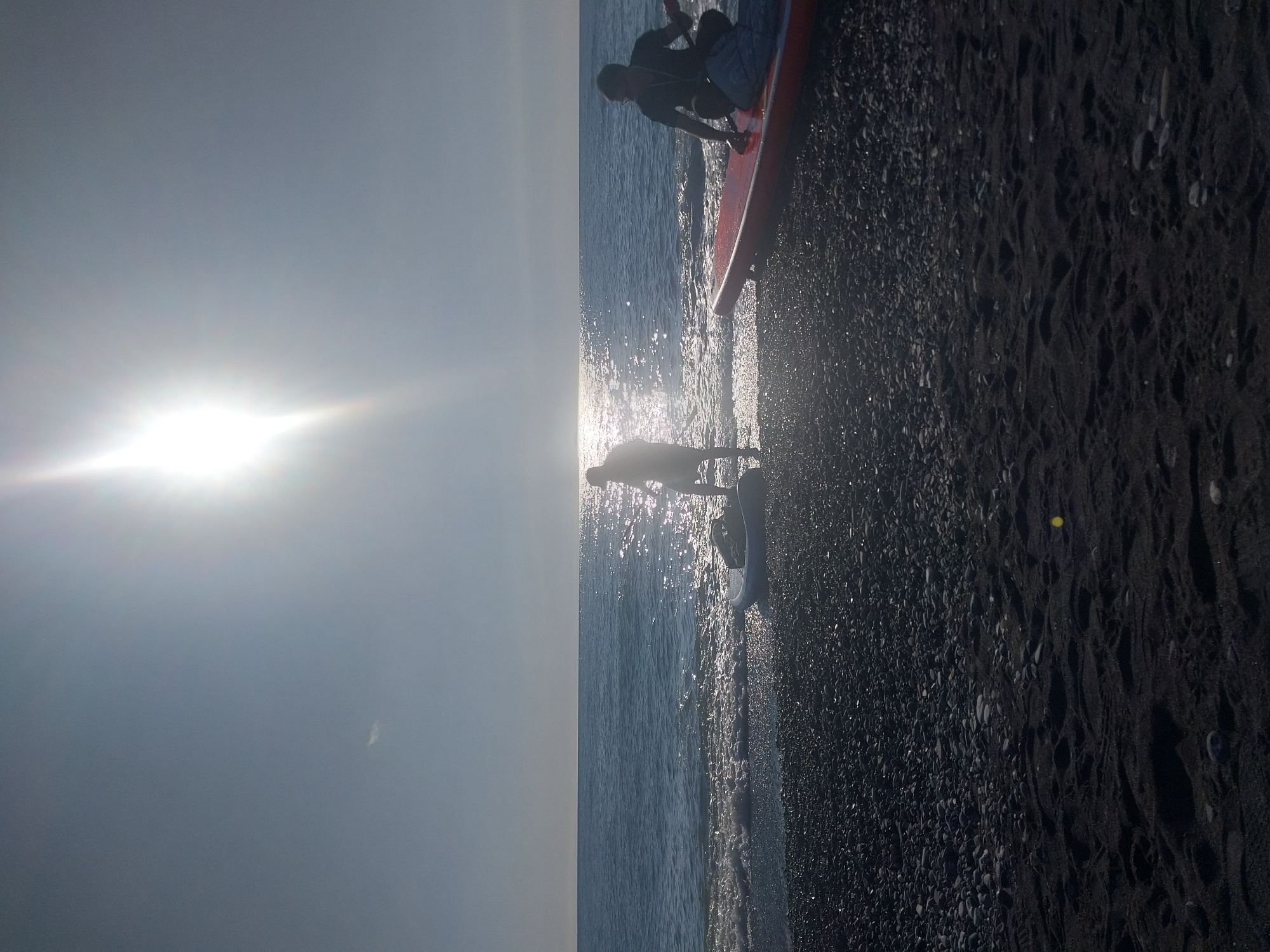 The image captures a sunlit beach scene, viewed from a slightly elevated perspective. The main subjects are two silhouetted figures and an orange paddleboard or surfboat at the water's edge.
One figure stands in the shallow water near the shore, their body a dark outline against the bright reflections on the water. A second person is visible further back, crouching or bending over a vibrant orange paddleboard, which is partially submerged in the water and partly resting on the dark, pebbly beach. The board has visible white or light-colored straps or gear on its surface.
The setting is a beach with dark-colored sand or small pebbles, which contrasts with the sparkling water. The ocean stretches horizontally across the middle of the frame, with small waves breaking gently at the shore. A very bright sun is positioned on the left side of the image, casting a strong glare and long reflections across the water, illuminating the scene from behind the subjects and creating dramatic backlighting.
The activity taking place appears to be paddleboarding or a similar water sport, with the individuals either preparing to launch or having just returned. The strong backlighting and the low angle of the sun suggest either early morning or late afternoon. The weather is clear and sunny. No visible text is present in the image.