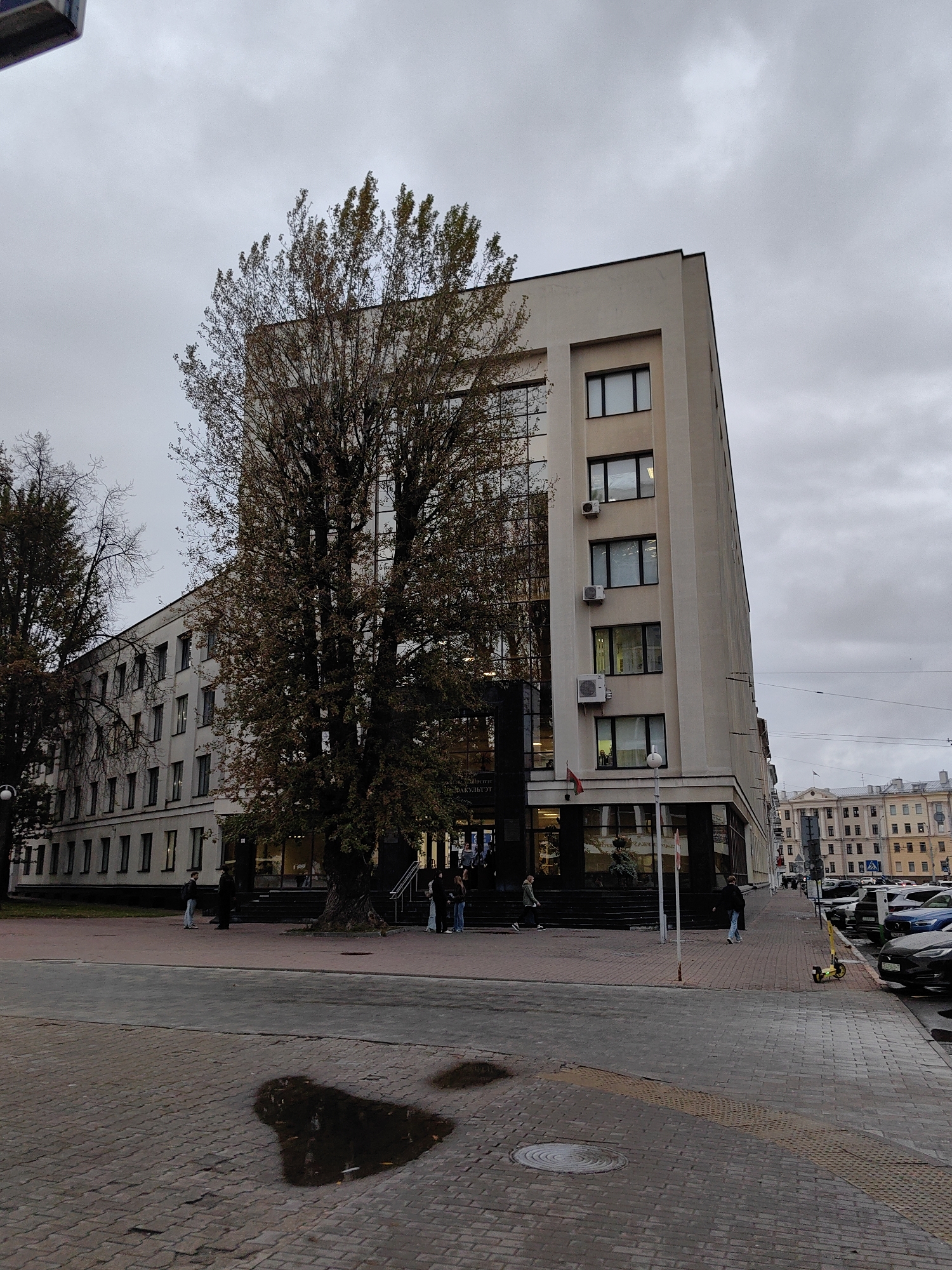 The image depicts a street view in Minsk, Belarus, under an overcast sky. A large, multi-story building with beige and glass facades dominates the right side of the frame. Adjacent to it, a longer, white building with numerous windows stretches across the left. A large tree with sparse autumn foliage stands prominently in front of the beige building.
On the street, several people are visible. Two individuals stand near the entrance of the beige building, and a few others walk along the sidewalk. A yellow electric scooter is parked on the sidewalk. Cars are parked along the street on the right side of the image. The foreground shows a cobblestone pavement with a large puddle reflecting the sky. In the distance, other buildings can be seen, suggesting an urban environment.
The overall impression is a somewhat muted urban scene on a cloudy day.