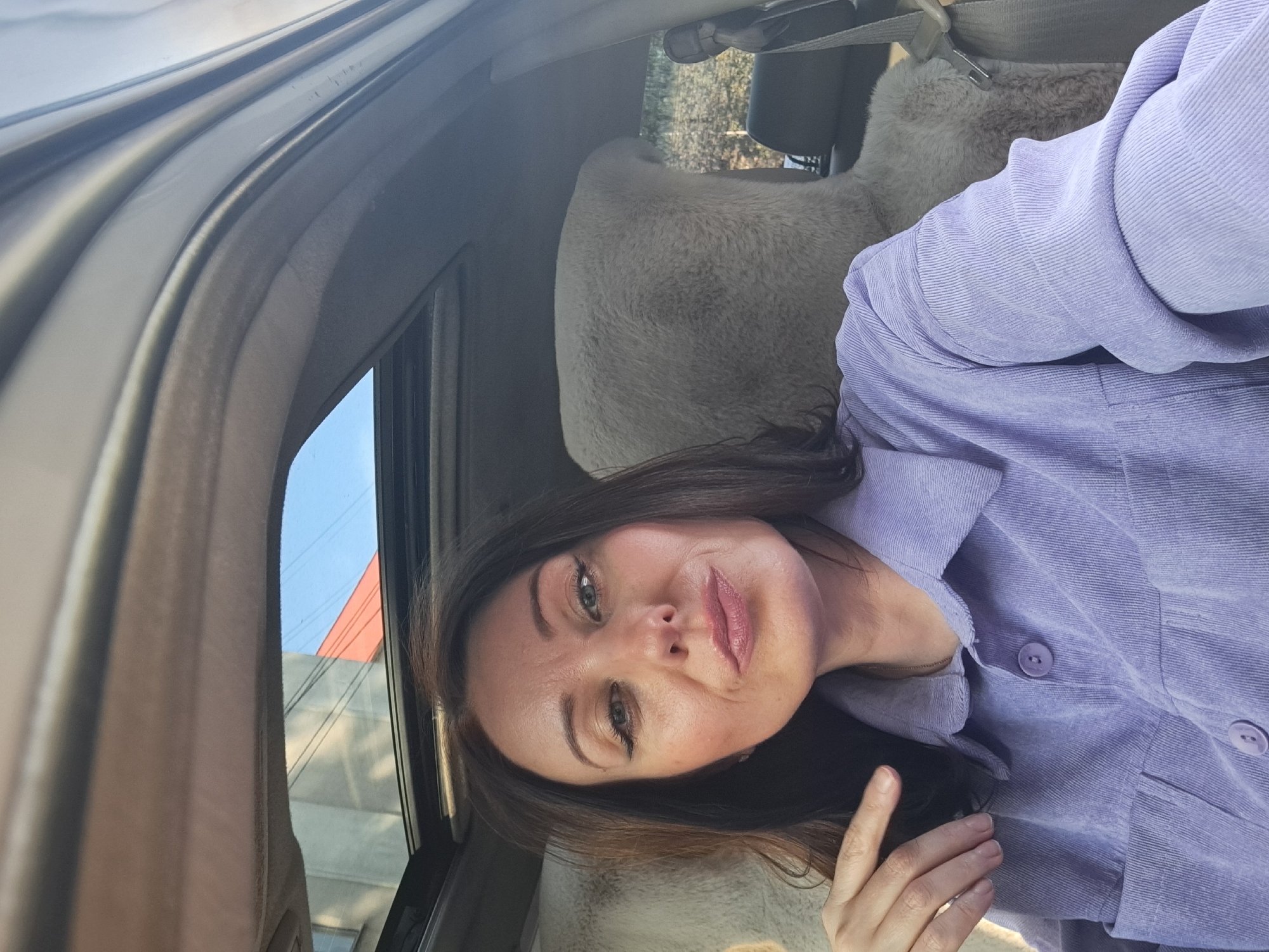 A medium shot captures a woman inside a vehicle, seated in the driver's seat. She has shoulder-length dark brown hair and is wearing a light purple corduroy shirt. Her left hand is resting on her chest, near her collarbone. The vehicle's sunroof is open, revealing a blue sky and the facade of a building with an orange roof. The interior of the car is visible, including plush, light-colored seat covers. The image appears to be a selfie, taken by the woman herself. The background suggests an urban environment, possibly Irkutsk, Russia, given the architectural style visible through the sunroof. The lighting indicates it is daytime.