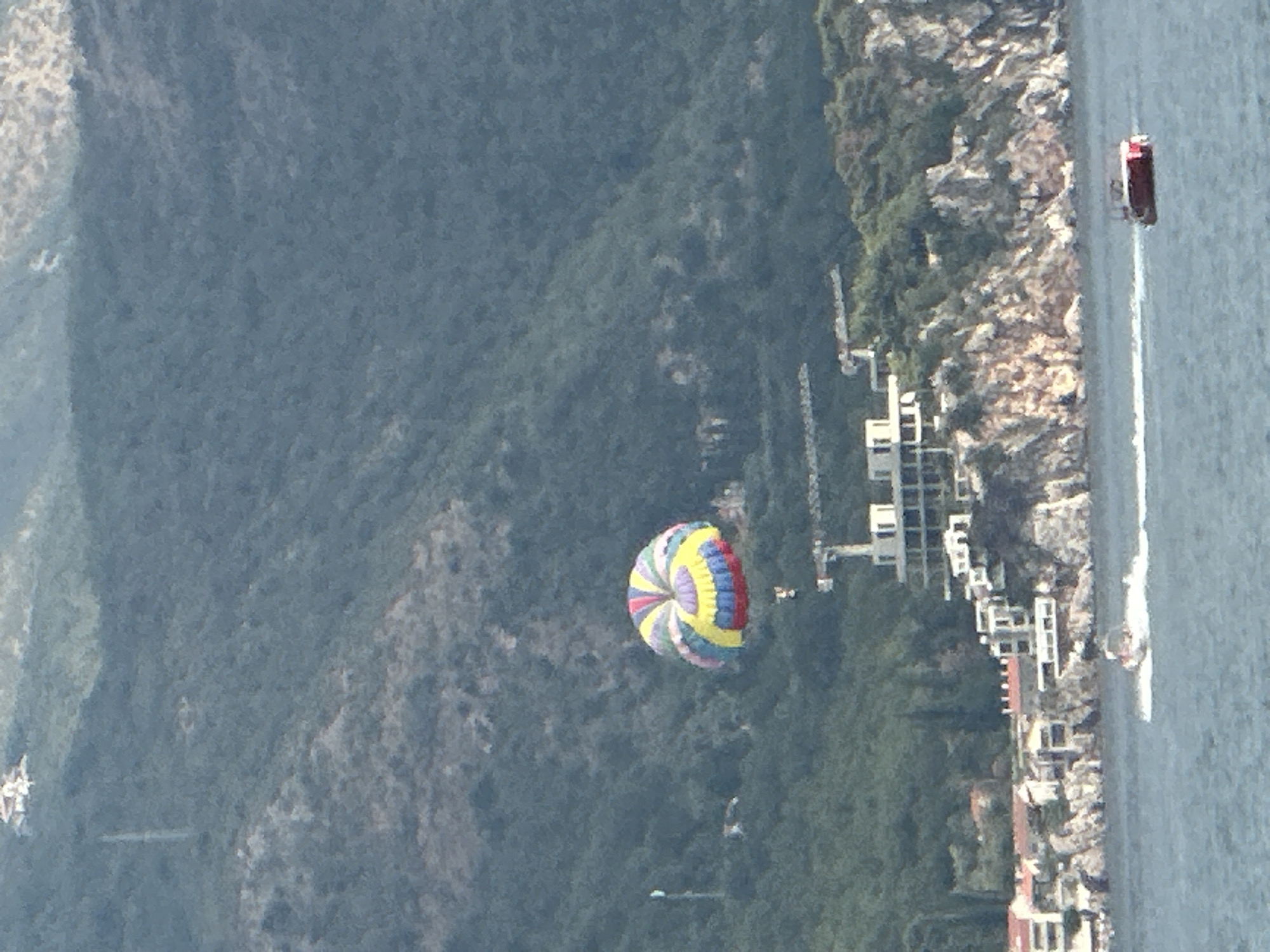 A parasailer with a multi-colored parachute is airborne over the water. The parachute is yellow, blue, red, and white. The parasailer is suspended below the parachute. In the background, a hillside is covered in green trees. A building complex is visible on the hillside, featuring white structures and cranes. The sea is visible in the foreground with a white boat leaving a wake. The location is near the city of Boreti, Montenegro.