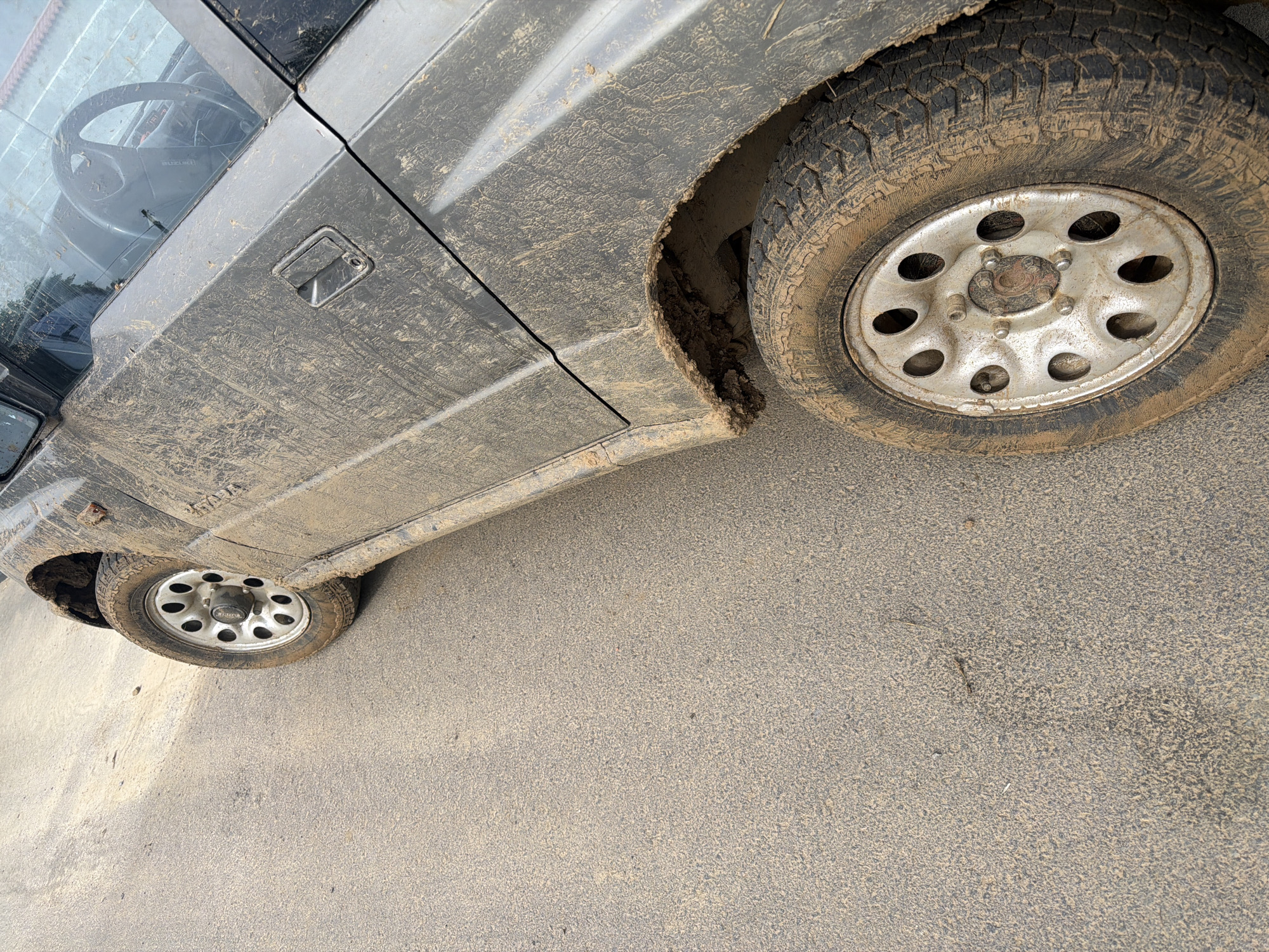 The image captures a close-up, angled view of a heavily mud-splattered dark-colored vehicle, likely an SUV or off-road model, situated on a dusty or sandy road in Malomir, Bulgaria. The vehicle's side, extending from the front door to the rear wheel, is almost entirely caked in dried light brown mud and dirt, indicating recent travel through challenging terrain.
The visible subjects include the vehicle itself and the road surface. Two wheels are partially visible: a rear wheel dominating the right side of the frame and a front wheel glimpsed at the bottom left. Both wheels are fitted with robust, knobby all-terrain tires, which are also heavily soiled with mud. The silver-colored metal rims feature multiple circular cutouts. A thick clump of mud is notably dislodged and hanging from the rear wheel arch, suggesting the force of movement through very wet conditions.
The car's door and door handle are coated in mud, though the handle's shape is still discernible. Through the car's window, a black steering wheel is visible, on which the text "SUZUKI" can be clearly read, identifying the vehicle's manufacturer. The ground beneath and around the vehicle is a light brown, granular surface, appearing dusty or sandy, consistent with the mud covering the car.
The scene depicts the aftermath or current state of a vehicle that has been used off-road. The bright, even lighting suggests it is daytime. No people are visible, and no specific activity beyond the vehicle's state is ongoing. The overall impression is one of rugged use in an outdoor, possibly rural, setting.