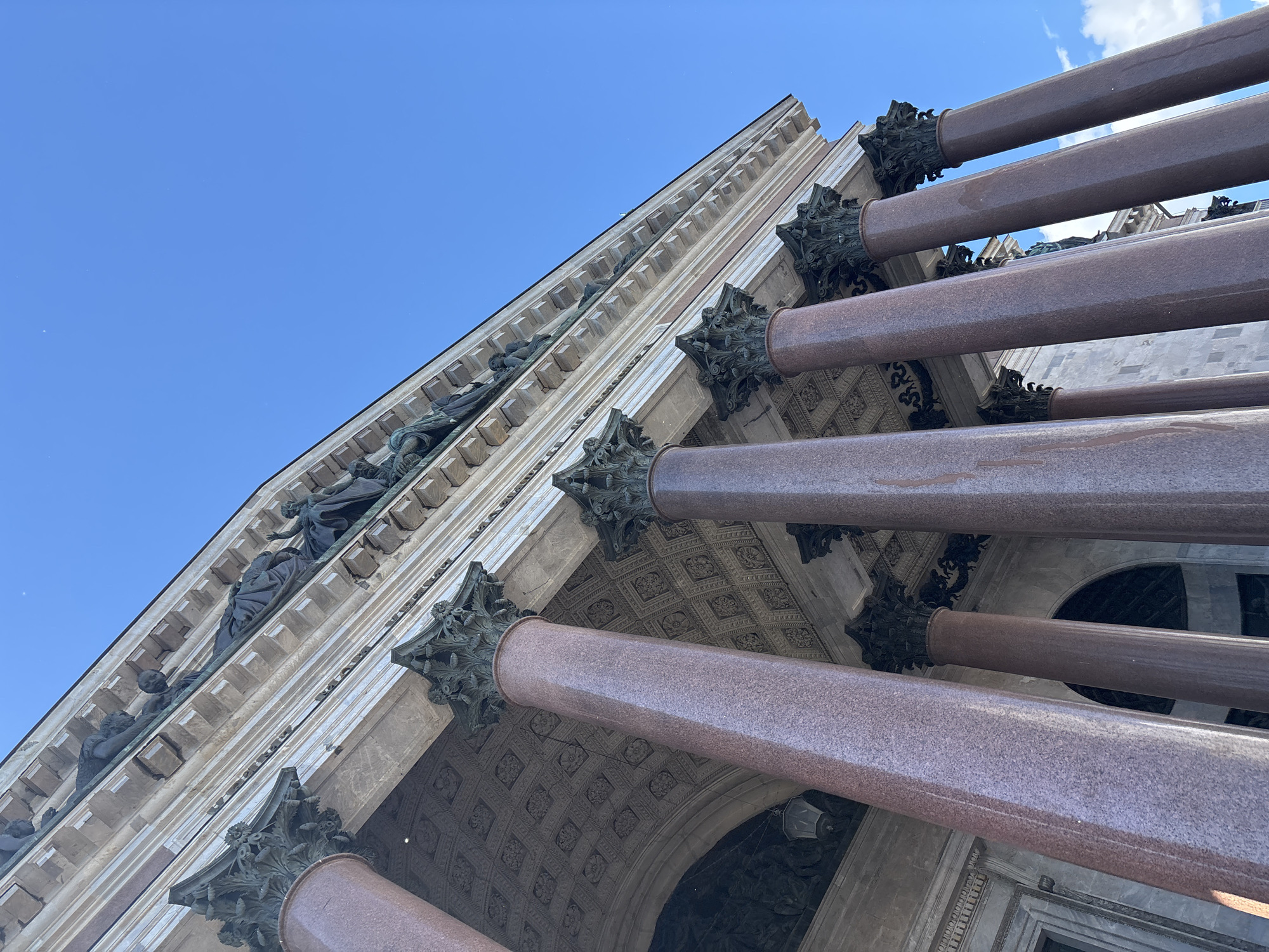 The image captures a low-angle, wide shot of the exterior of a grand building, likely a historical monument or cathedral, set against a clear blue sky with a few wispy clouds. The architecture is characterized by massive, ornate columns made of reddish-pink granite, interspersed with intricate stonework and sculptures. These columns are positioned in a way that suggests they are part of a colonnade or portico.
Visible on the facade of the building are detailed sculptures of human figures, appearing to be in various poses, adding a sense of narrative and historical depth to the structure. The stonework features repetitive, decorative patterns, particularly on the ceiling visible between the columns. The lighting suggests it is daytime, with the sun casting shadows and highlighting the textures of the stone and granite. The overall impression is one of grandeur, historical significance, and impressive architectural achievement. The clear sky and bright light indicate fair weather, likely a pleasant day for visiting such a landmark. The perspective, looking upwards, emphasizes the monumental scale of the building. No visible text is present in the image.