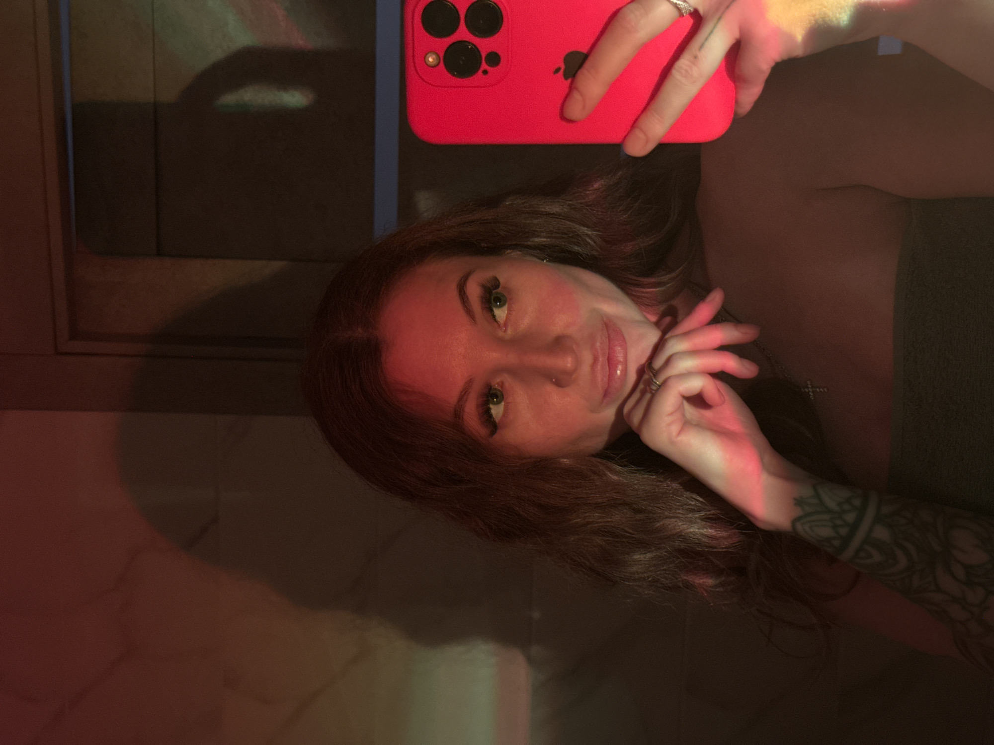 A selfie captures a woman taking a photo with a bright pink iPhone, likely in Chaikovsky, Russia. She has long, dark brown hair and striking green eyes, enhanced with makeup, and wears a nose piercing. Her expression is calm with a slight smile. The woman holds the iPhone in her right hand, while her left hand rests gracefully near her chin. She is adorned with rings on her fingers and a visible silver cross necklace. A detailed sleeve tattoo covers her left arm. The background is dark and indistinct, suggesting an indoor setting like a room, possibly at night or in the evening. Atmospheric, colorful lighting, featuring prominent red, orange, and green hues, casts reflections across her face, hands, and the surrounding surfaces, creating a moody ambiance. The Apple logo is clearly visible on the back of the bright pink iPhone.