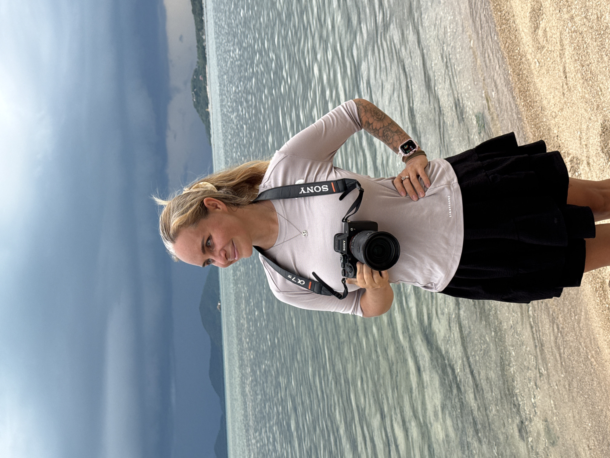 A woman with blonde hair pulled back in a ponytail stands on a sandy beach, holding a Sony camera with a strap around her neck. She is wearing a light-colored t-shirt and a black skirt. Her left hand is on her hip, and her right hand is holding the camera. In the background, there is a calm body of water with a distant shoreline featuring hills and trees under a cloudy sky. The scene is set in Ban Bang Khao, Thailand.