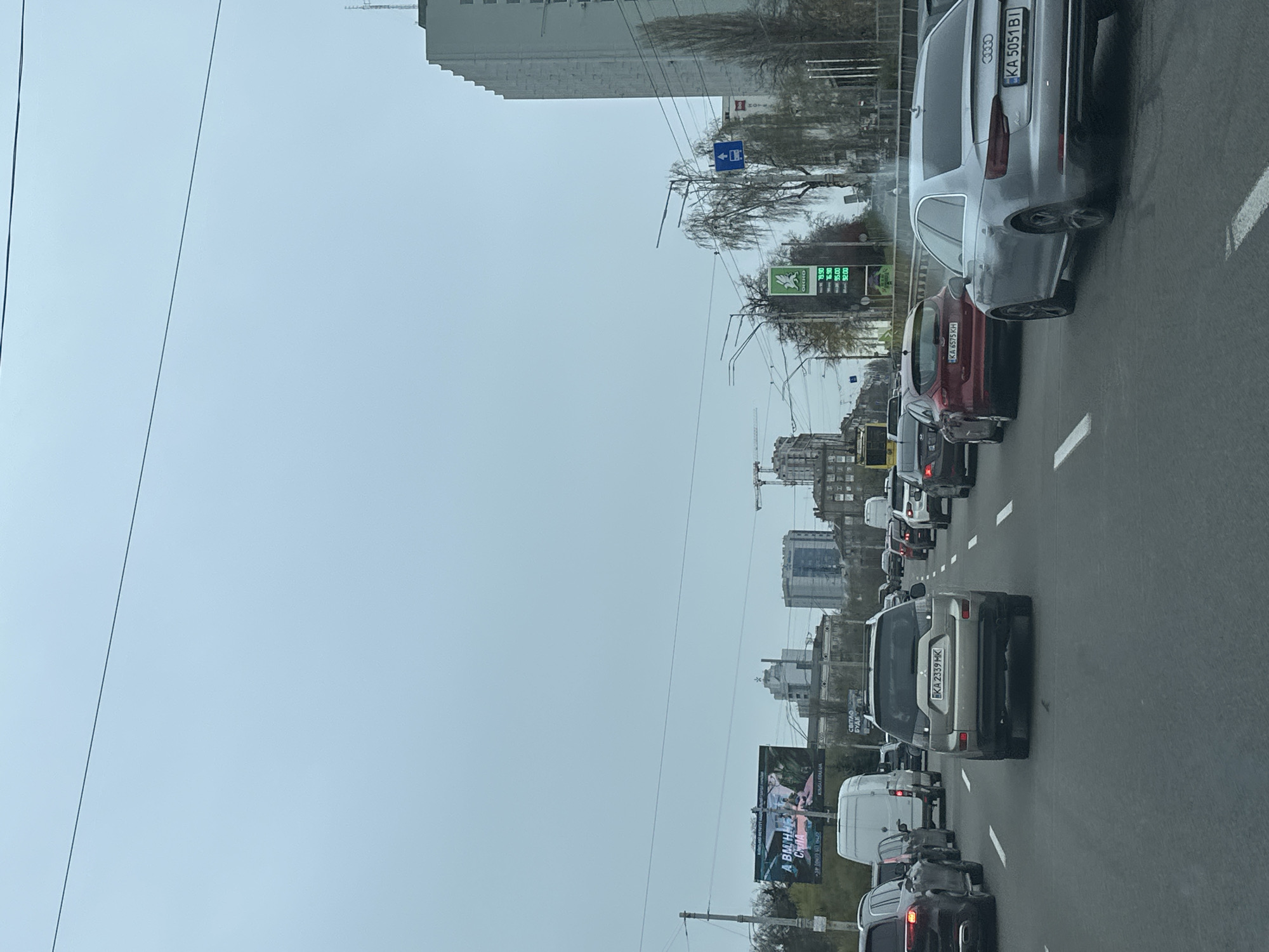 The image shows a congested road in Kyiv, Ukraine, likely due to traffic. The camera's point of view is from within a vehicle on the road. There are several vehicles in view. A silver Audi's license plate reads "KA 5051 BI." A red car behind the Audi has the license plate "KA 6575 KH." There is a silver Mitsubishi SUV in front of them with the license plate "EKA 2339 MK." Billboards are visible on the left. A gas station sign is visible in the background with the brand name "OKKO". The sky is overcast and gray. Buildings are seen on the left side, and there is a construction crane in the distance. Power lines are overhead.