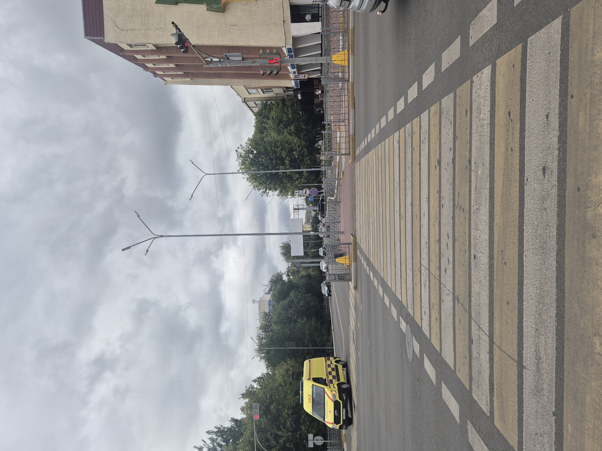 The image depicts a street intersection in Kokshetau, Kazakhstan, under a cloudy sky. A yellow ambulance with the number "103" on its side is driving through the intersection from left to right. A pedestrian crossing with yellow and white stripes is prominent in the foreground, leading across the asphalt road. On the right side of the road, a multi-story building with a beige and brown facade is visible, featuring architectural details and windows. A traffic light displaying a red signal is mounted on a pole in front of the building. Streetlights and trees line the sides of the road, contributing to the urban environment. In the background, other vehicles and buildings are partially visible. The scene appears to be during daytime, with overcast weather conditions.