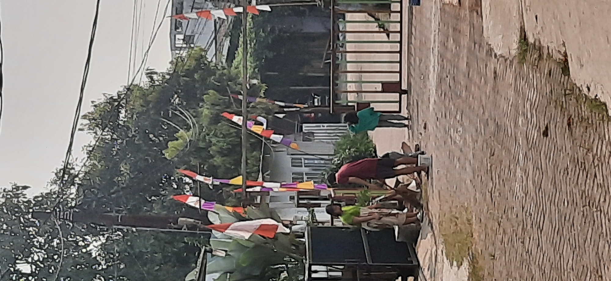In Cisauk, Indonesia, an outdoor scene captures a glimpse of daily life with a festive touch. A paved area, possibly a street or courtyard, features a brick pattern. In the background, a building with white walls and dark-trimmed windows stands amidst lush green trees. Overhead, power lines stretch across the sky, supporting numerous Indonesian flags, which are red and white, strung between poles and trees, adding a vibrant, patriotic flair.
In the foreground, a group of people is engaged in various activities. A person in a green shirt and patterned pants stands near a dark, metal object, possibly a cabinet or enclosure. They are facing another individual in a red shirt and dark shorts. This second person appears to be attending to something on the ground, perhaps an animal or objects, with a white bucket nearby. To the right, a child in a turquoise hoodie and jeans stands near a colorful metal gate with vertical bars. The gate is closed, separating the paved area from what lies beyond. The overall impression is one of a community gathering or a moment of everyday interaction, amplified by the presence of national flags.
