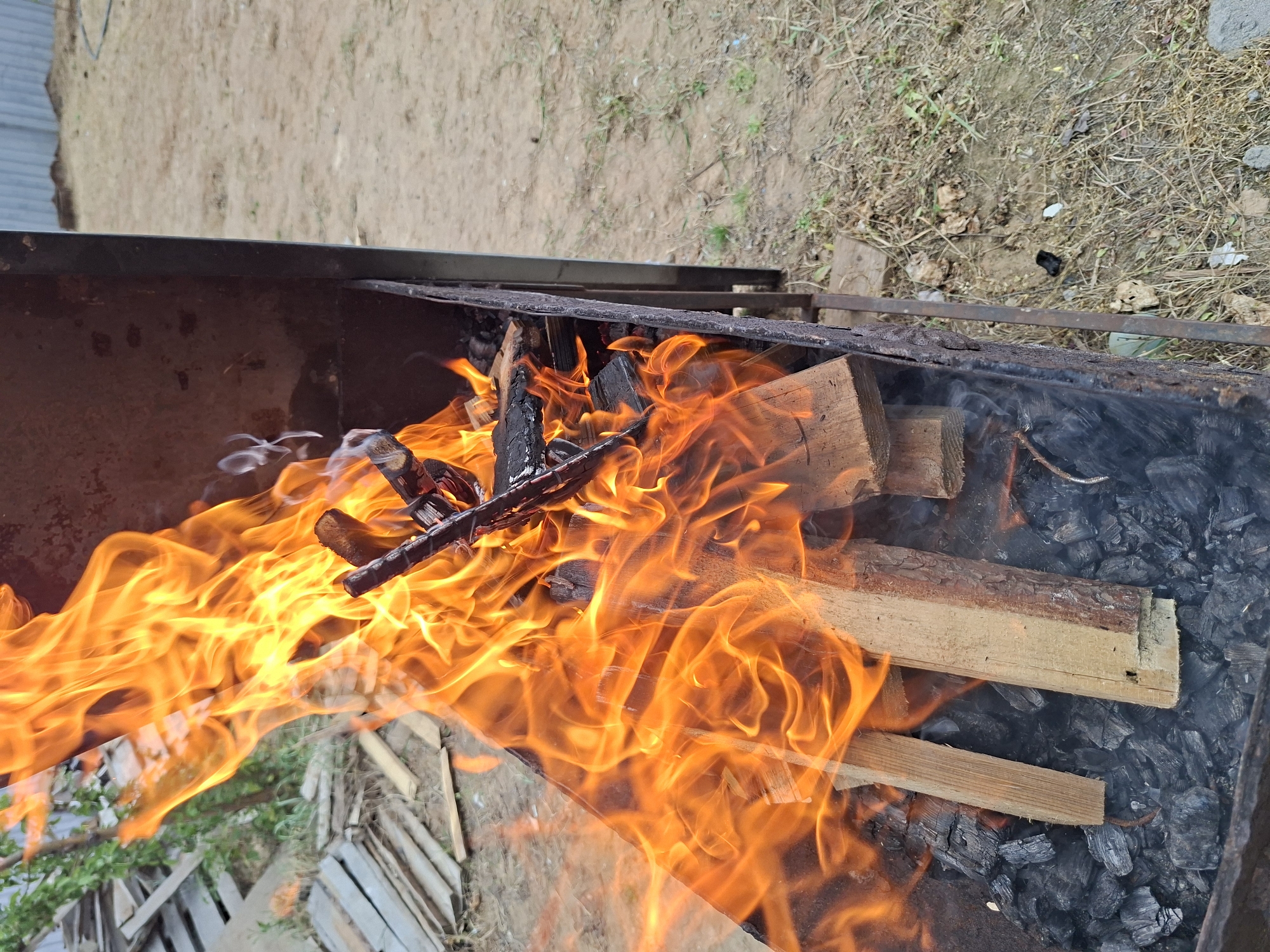 The image shows a metal barbecue grill filled with burning wood and charcoal. The fire is intense, with bright orange flames engulfing the wood. Some smoke is also visible. The grill is located outdoors, possibly in a backyard or park, as indicated by the ground and vegetation visible in the background. The location is Unknown, Unknown.