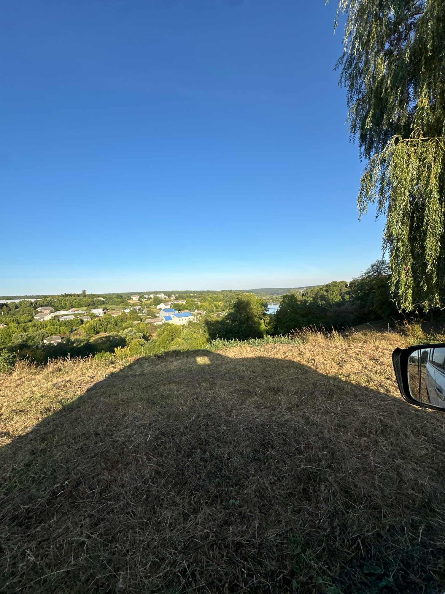 The image shows a wide, panoramic view from a grassy hill overlooking a village in Makiv, Ukraine. The sky is a clear, bright blue with no clouds. On the right side of the frame, the drooping branches of a willow tree hang down, partially obscuring the view. In the foreground, the dry, yellowed grass covers the hill. A dark shadow, likely cast by a vehicle, covers a significant portion of the foreground. A side-view mirror of a white vehicle is visible on the far right edge of the image, reflecting a portion of the scene. In the mid-ground, a village is nestled among lush green trees. Houses with various colored roofs, some blue, are scattered throughout the village. A body of water, possibly a river or lake, can be seen in the distance beyond the village. The overall impression is one of a peaceful rural landscape on a sunny day.