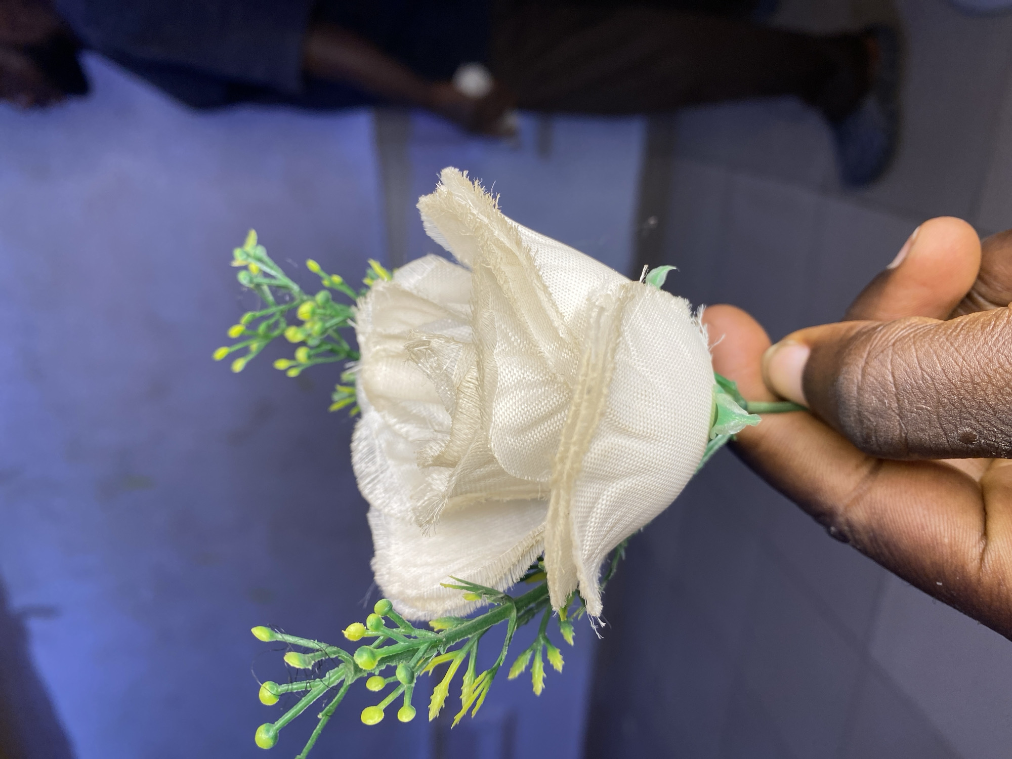 A hand holds a white, faux rose with green and yellow accent foliage. The hand appears to be dark-skinned. The rose is made of fabric with frayed edges. The background is blurry and appears to show a room with other people present. There is no visible information in the image to ascertain that the photo was taken in Bukuru, Nigeria.