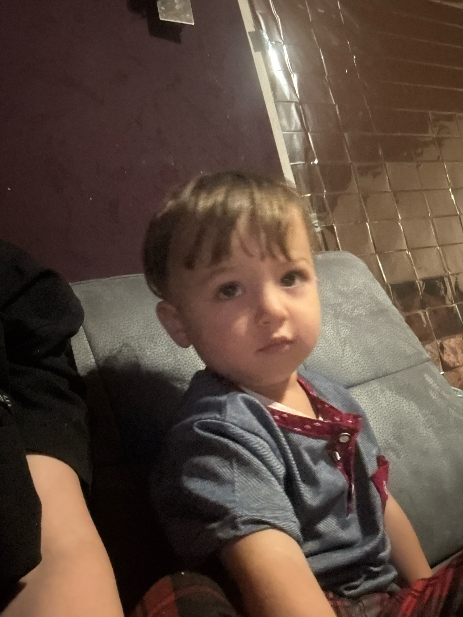 A young child with light brown hair sits on a gray couch. The child is wearing a blue collared shirt with a red trim and buttons, and red and black plaid pants. A person wearing a black hooded sweatshirt is seated to the left of the child, with their arm visible. The background consists of a dark purple wall and a wall covered in reflective, square tiles that appear to be made of foil. The scene is indoors. The location is the city of Überherrn, Germany.