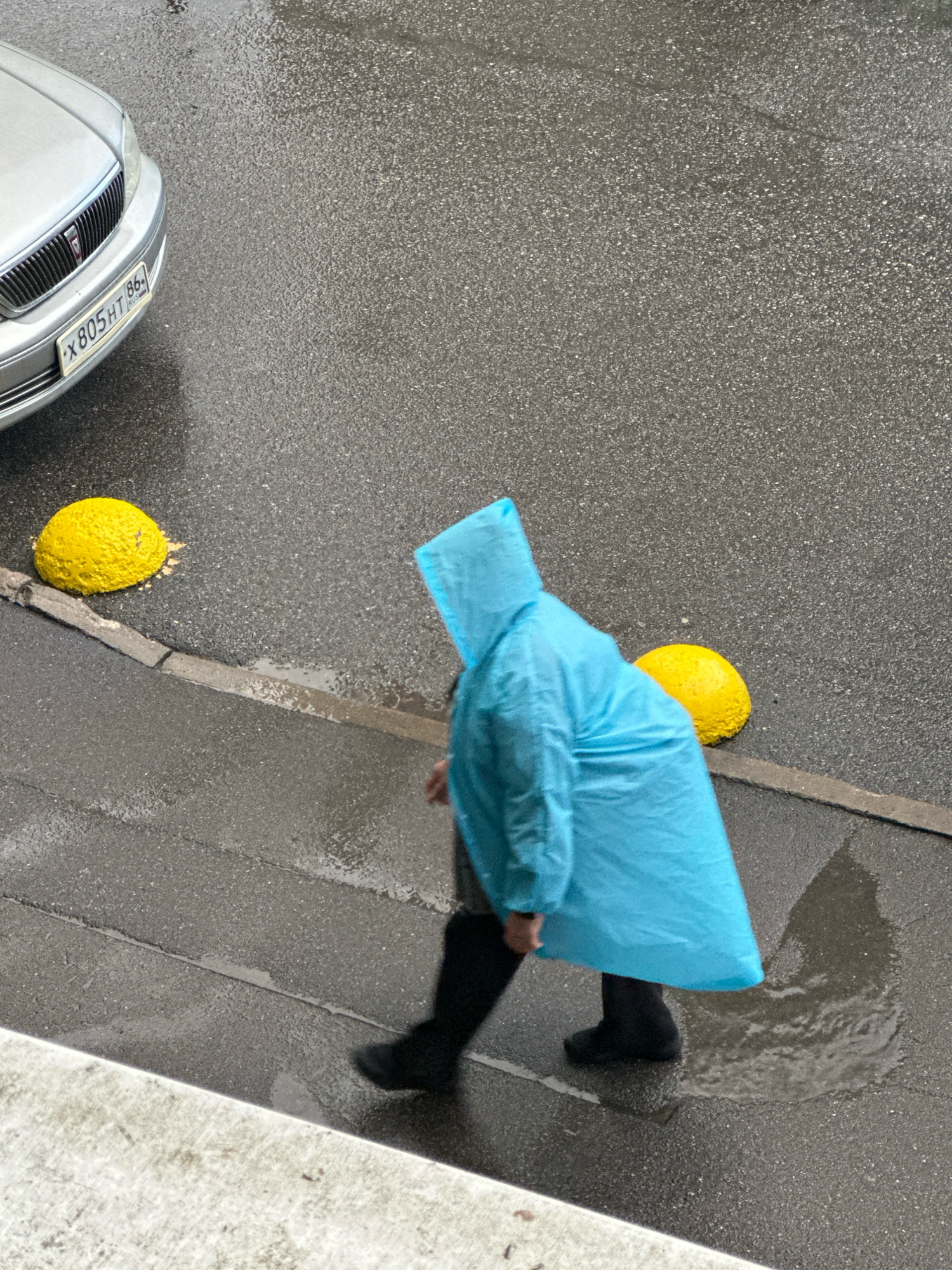 An overhead, medium-distance shot depicts a person walking on a wet asphalt surface in what appears to be an urban environment. The individual, clad in a bright blue disposable poncho, is the central focus, their form largely obscured by the voluminous garment. The poncho's hood is up, and the person's lower legs are visible, clad in black trousers and dark shoes, stepping through a shallow puddle.
To the left of the frame, the front quarter of a silver sedan is visible, its license plate reading "X805HT 86 RUS". The car is parked on the wet asphalt, and its headlights are on.
Two bright yellow, rounded concrete bollards are positioned along the edge of the asphalt, one to the left of the person and slightly ahead, and another to the right and further back. The ground is visibly wet, with reflections of the bollards and the car visible on the surface, indicating recent rainfall or damp conditions. A concrete curb separates the asphalt from a lighter-colored pedestrian walkway or building facade in the foreground. The overall scene suggests a rainy day in a city, possibly in Russia given the license plate.