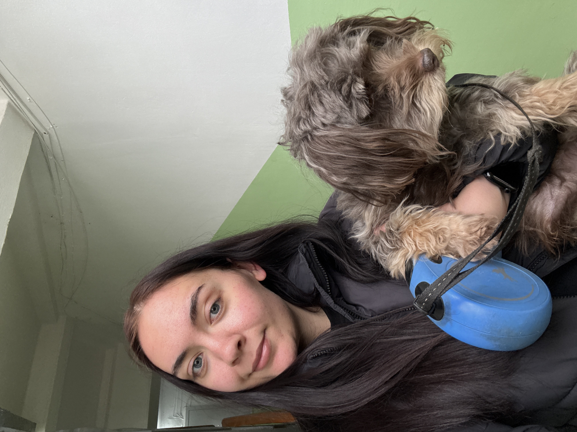 A young woman with long dark hair and light blue eyes is posing for a selfie with a small, shaggy brown and grey dog. The dog is wearing a black jacket. The woman is wearing a black jacket, and a blue retractable leash is visible. The background consists of a wall painted white and green. It is not possible to determine the location as Chaikovsky, Russia from the given image.