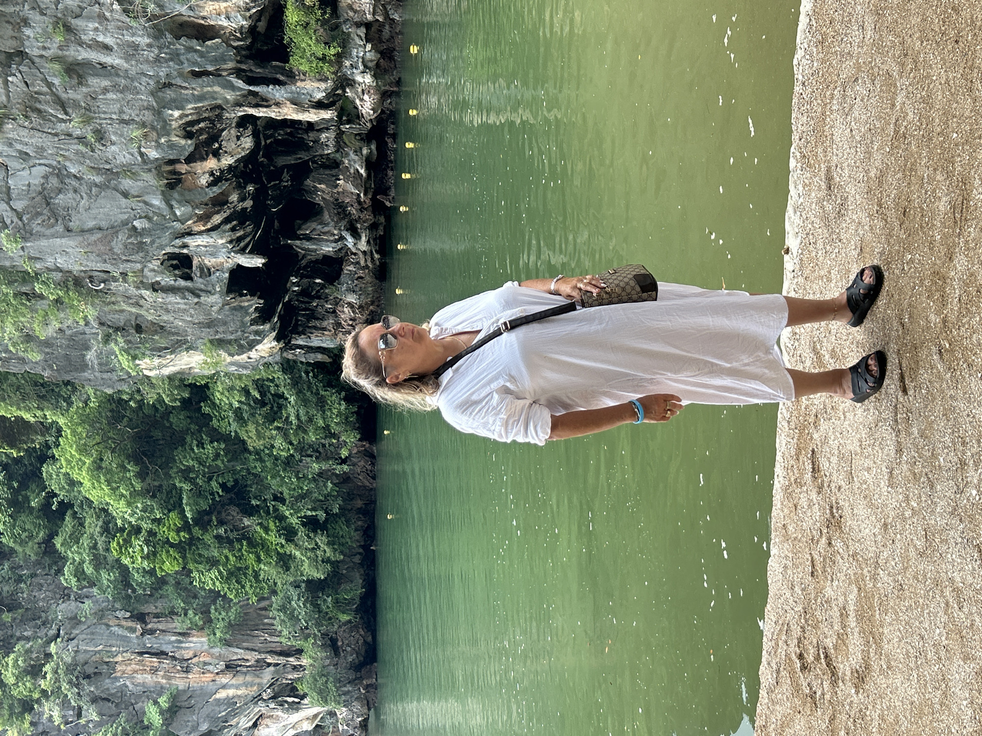 The image shows a woman standing on a sandy shore next to calm, green water. She is wearing a long white dress, sunglasses, and black sandals. A small brown and beige patterned purse is slung across her body. She is looking down and to her right, with her left hand resting on her stomach and her right arm bent at the elbow. The background features a steep, rocky cliff face with lush green foliage growing on its upper slopes. Several small, circular lights are visible along the edge of the cliff face, reflected in the water. The water appears still with some white flecks floating on the surface. The lighting suggests it is daytime, with the sun casting shadows. The scene evokes a sense of peacefulness and natural beauty. The location is identified as Tambon Krasom, Thailand. There is no visible text in the image.