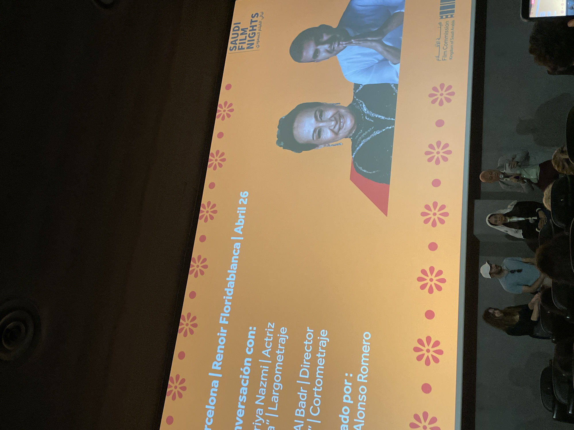 The image shows a presentation screen at a "Saudi Film Nights" event, likely taking place in Barcelona at the Renoir Floridablanca cinema on April 26th. The screen displays the event's title, logos of the Saudi Film Nights and Film Commission of Saudi Arabia, and the details of a conversation with actress Riya Nazmi about a feature film and director Al Badr about a short film, moderated by Alonso Romero. Two portraits of the actress and director are also on the screen.
In the foreground and to the right of the screen, a small section of the audience is visible seated in theater chairs, some identifiable individuals appearing to be engaged with the presentation. The environment appears to be a darkened cinema hall.