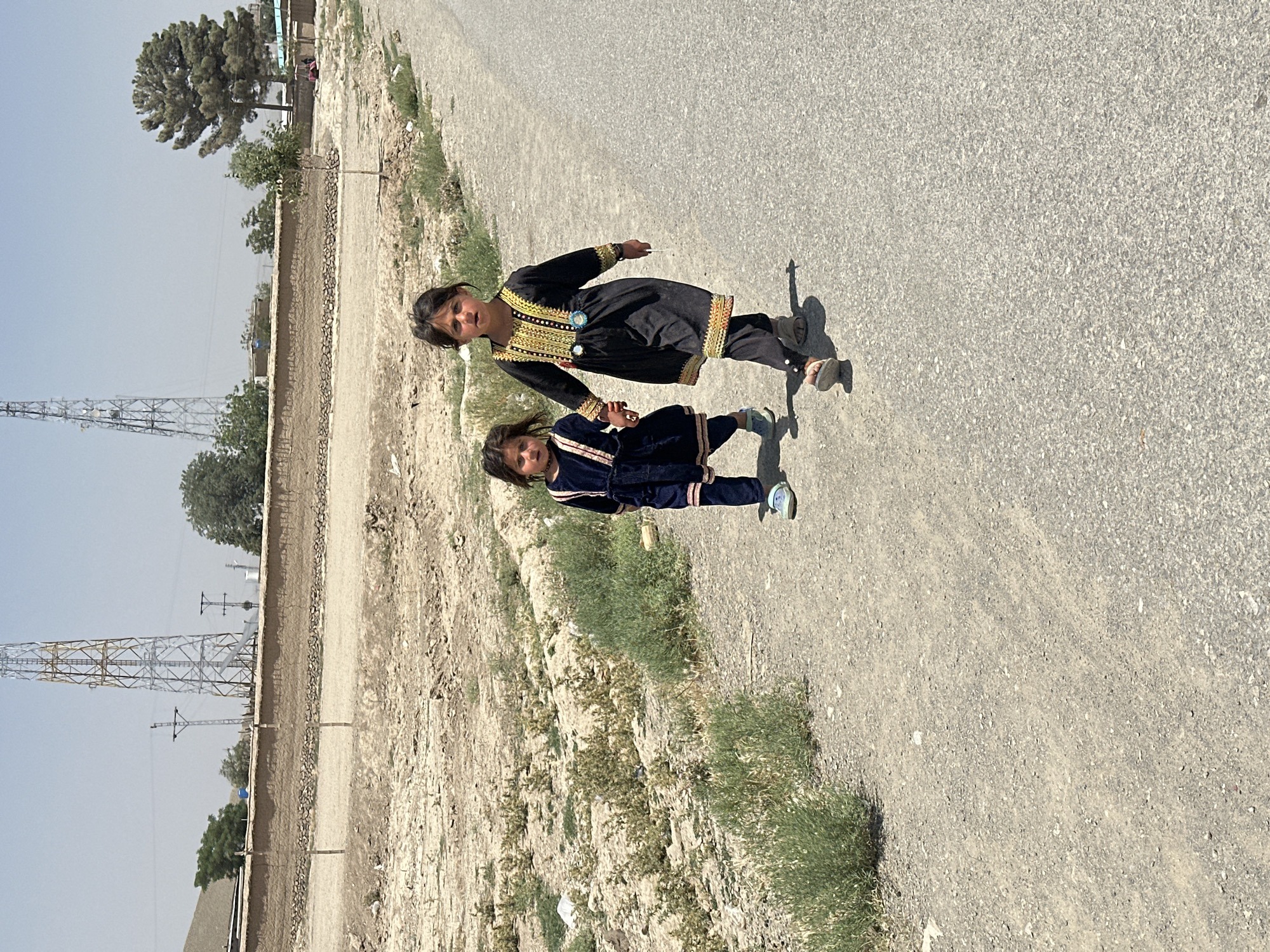 Two young girls, one older than the other, walk hand-in-hand along a paved road. The older girl, on the right, wears a black dress with gold and red embroidery and dark pants, and is looking towards the viewer. The younger girl, on the left, wears a dark blue dress and dark pants, and looks down as she walks. They are walking away from the camera and towards the viewer. The background consists of dry, dusty land with sparse vegetation, a low, mud-brick wall, and two large telecommunications towers in the distance. The sky is clear and the sun is bright, suggesting a warm day. The scene appears to be in an arid or semi-arid region, possibly within or near the city of Kanozai, Pakistan, given the presence of the towers which are common in such areas.