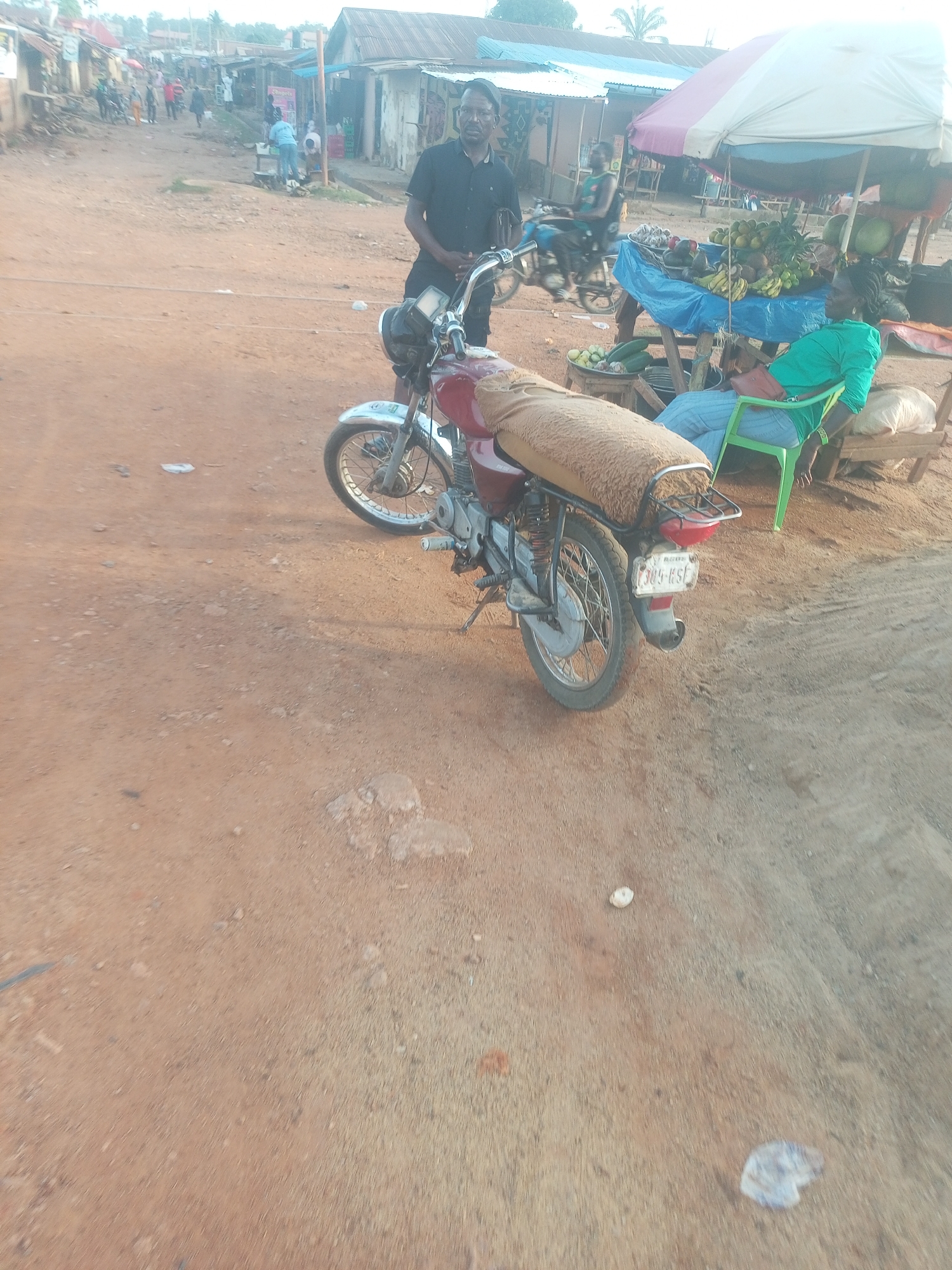 The image depicts a bustling street scene in a rural or semi-urban setting, consistent with Yelwa, Nigeria. The foreground and mid-ground are dominated by an unpaved, reddish-brown dirt road with scattered small rocks and debris.
In the mid-ground, a dark red motorcycle with a tan, fuzzy seat cover is parked, its front wheel facing left. A man in a black shirt and shorts stands beside the motorcycle, looking directly towards the camera, with his left hand resting on the handlebars.
To the right, a market stall operates under a striped umbrella (white and red/pink), showcasing a variety of fresh produce, including bananas, pineapples, watermelons, oranges, and other fruits and vegetables like cucumbers and limes. A woman, dressed in a green top and light blue pants, is seated in a vibrant green plastic chair next to the stall, leaning back and appearing relaxed or resting.
In the background, the dirt road continues uphill, lined with simple buildings, many featuring corrugated iron roofs. Several other people are visible walking or standing near stalls, indicating ongoing daily activities and commerce. The lighting suggests either early morning or late afternoon, with soft, warm illumination.
A notable detail is the motorcycle's license plate, which clearly displays "305-KSF". The overall scene captures a typical slice of daily life in a vibrant, informal outdoor market environment.